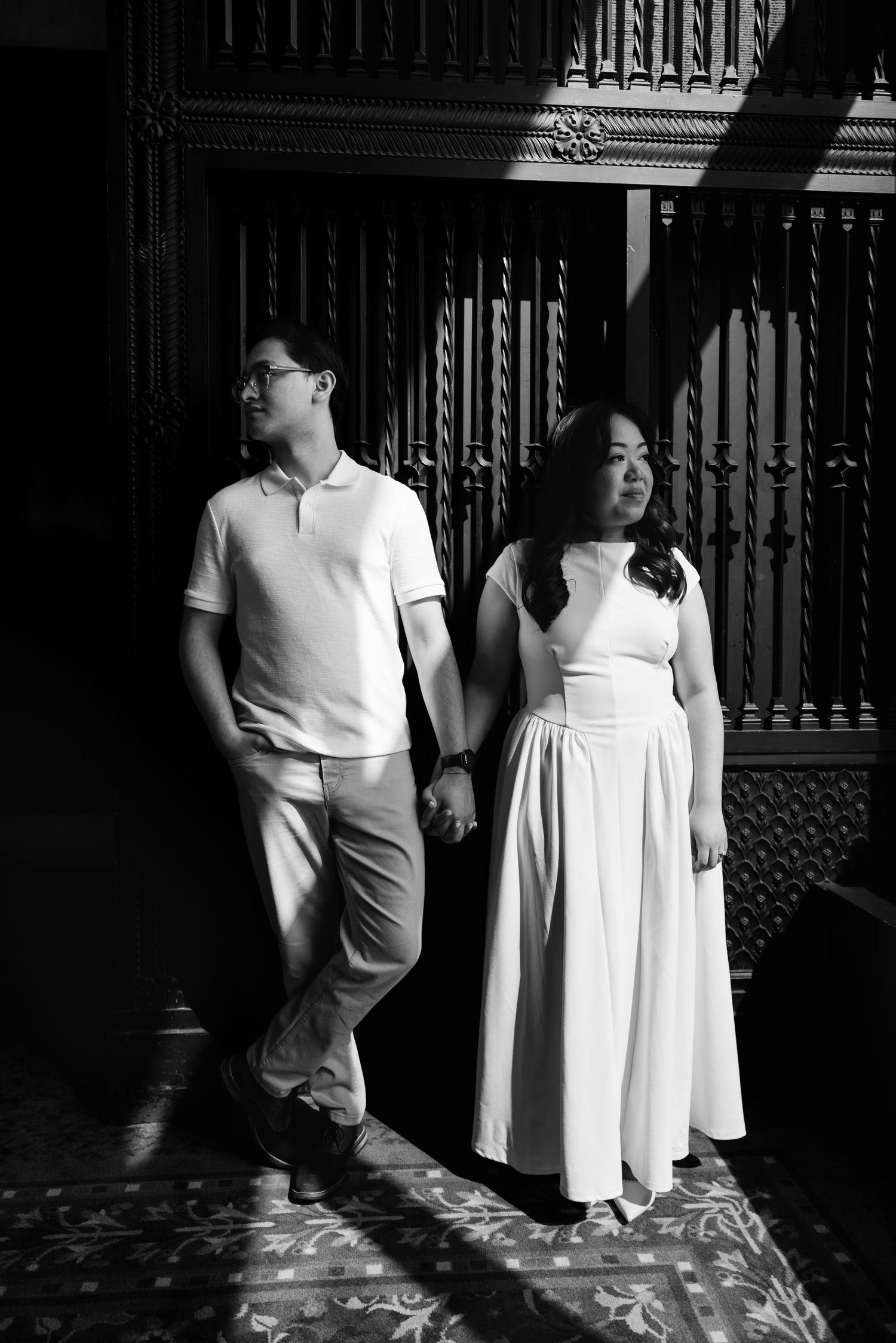 Black and white photo of couple holding hands standing before decorative wooden panels in grand interior.