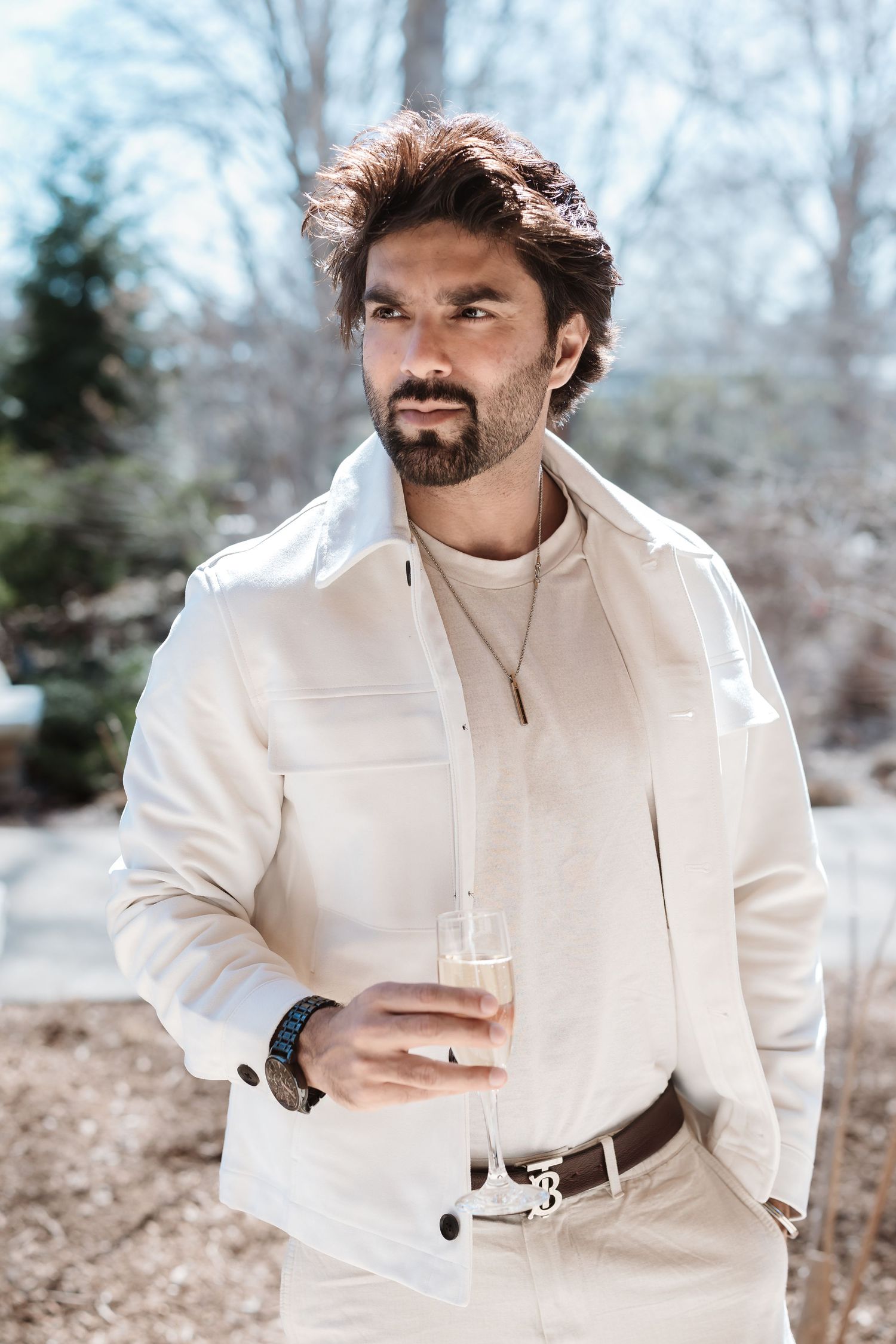 Stylish bearded man in white outfit holds champagne glass outdoors at winter event.
