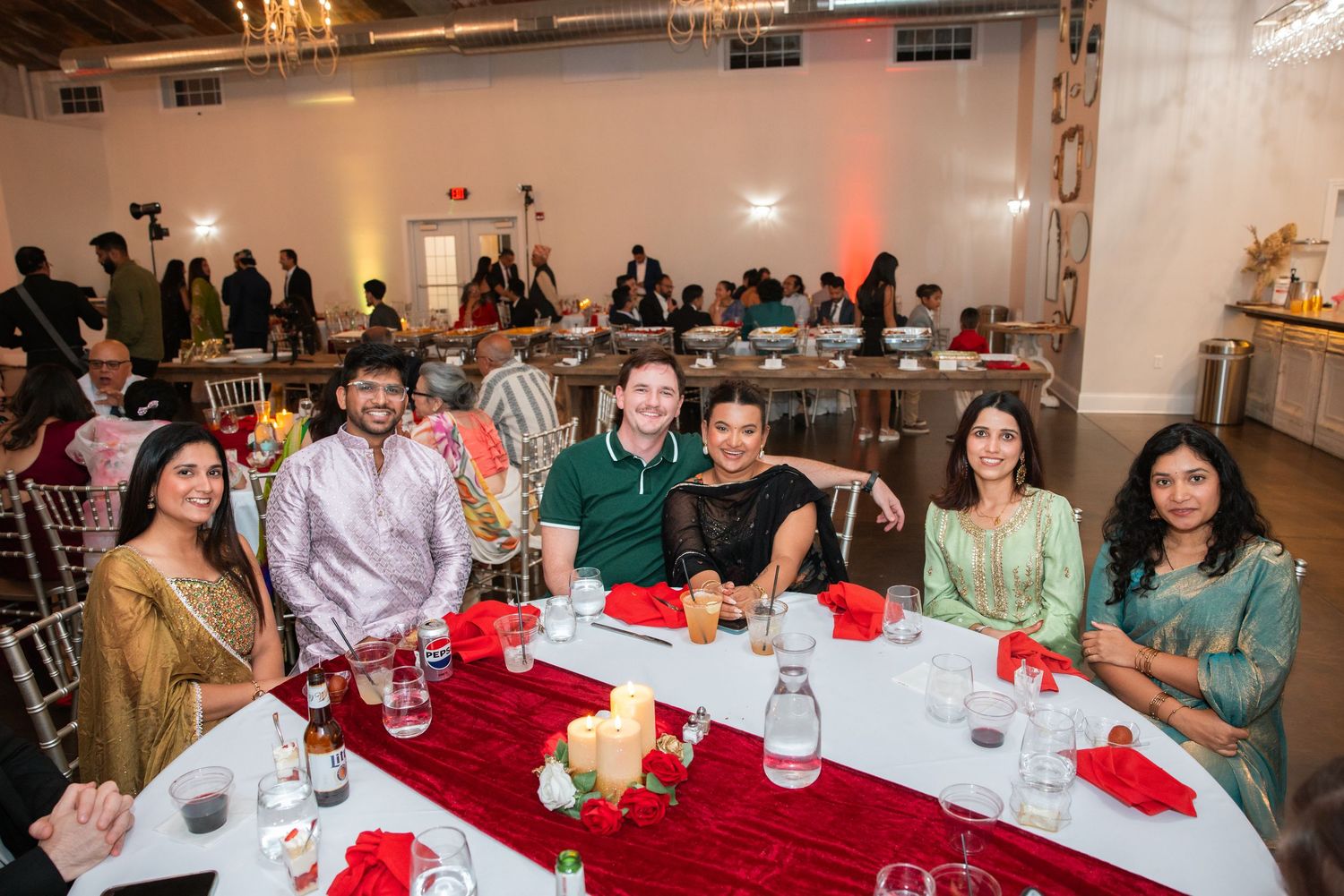 Six guests posing happily at a decorated banquet table during a vibrant Indian celebration.