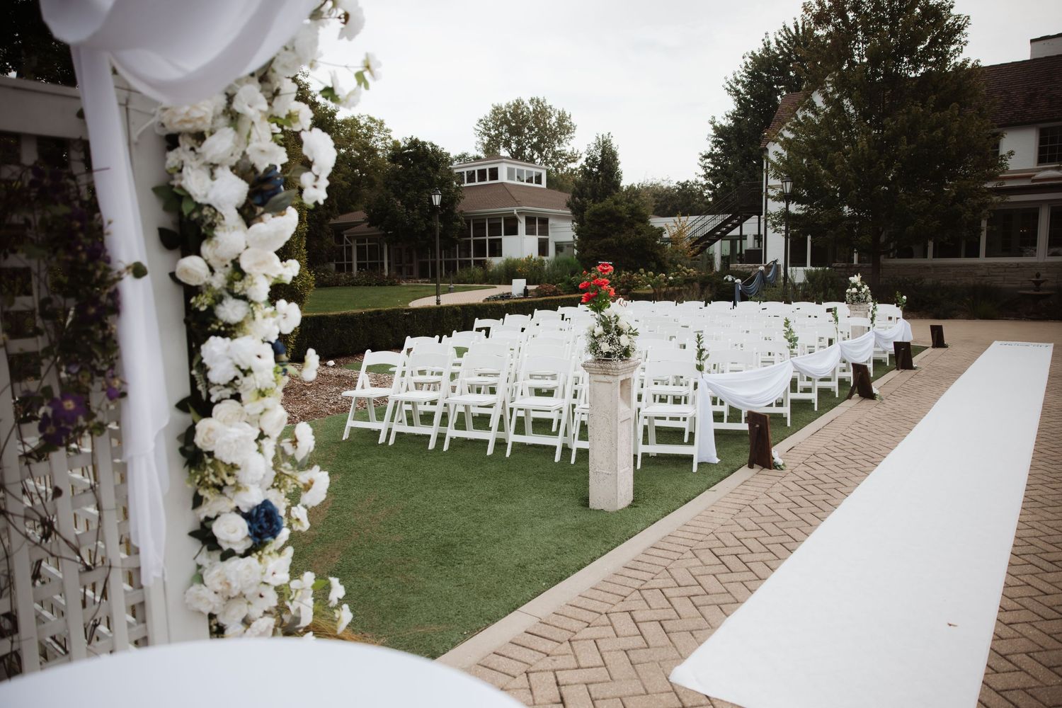 White floral-adorned wedding ceremony arch with white aisle runner and rows of white chairs on brick pathway.