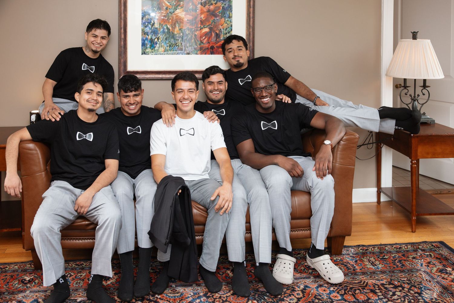 Happy groom surrounded by smiling groomsmen on leather couch in matching black shirts before wedding ceremony.