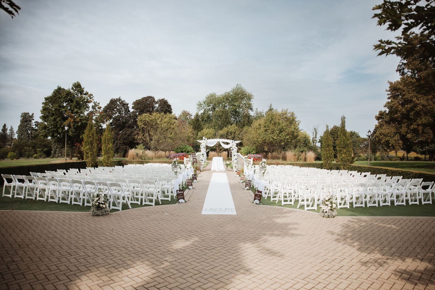 Elegant outdoor wedding ceremony with white folding chairs, floral arch, and brick paver aisle.