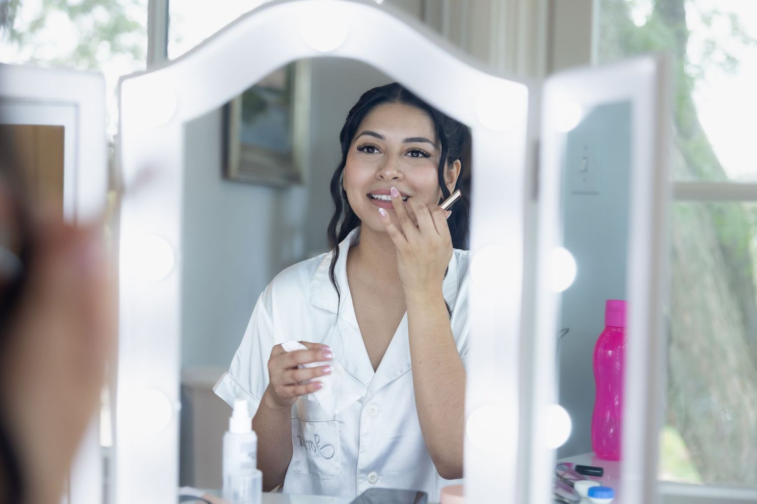 A young Asian woman applies makeup while looking at her reflection in a lighted vanity mirror.