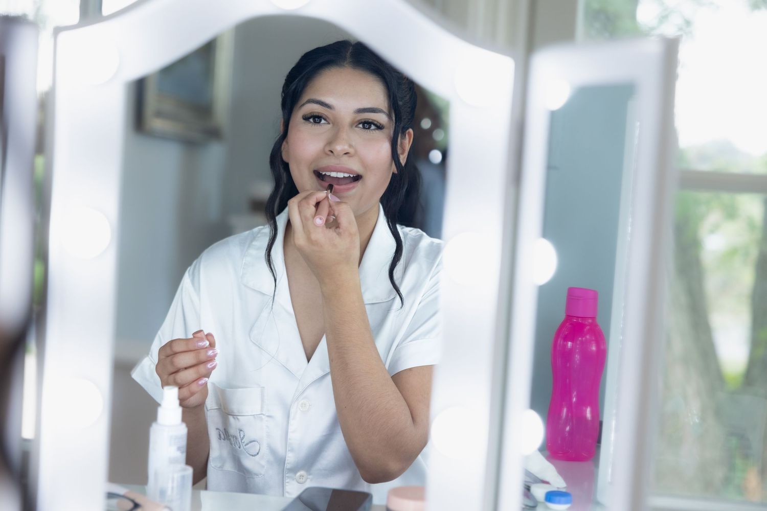 A young Asian woman applies lipstick while smiling at her reflection in a lighted vanity mirror.