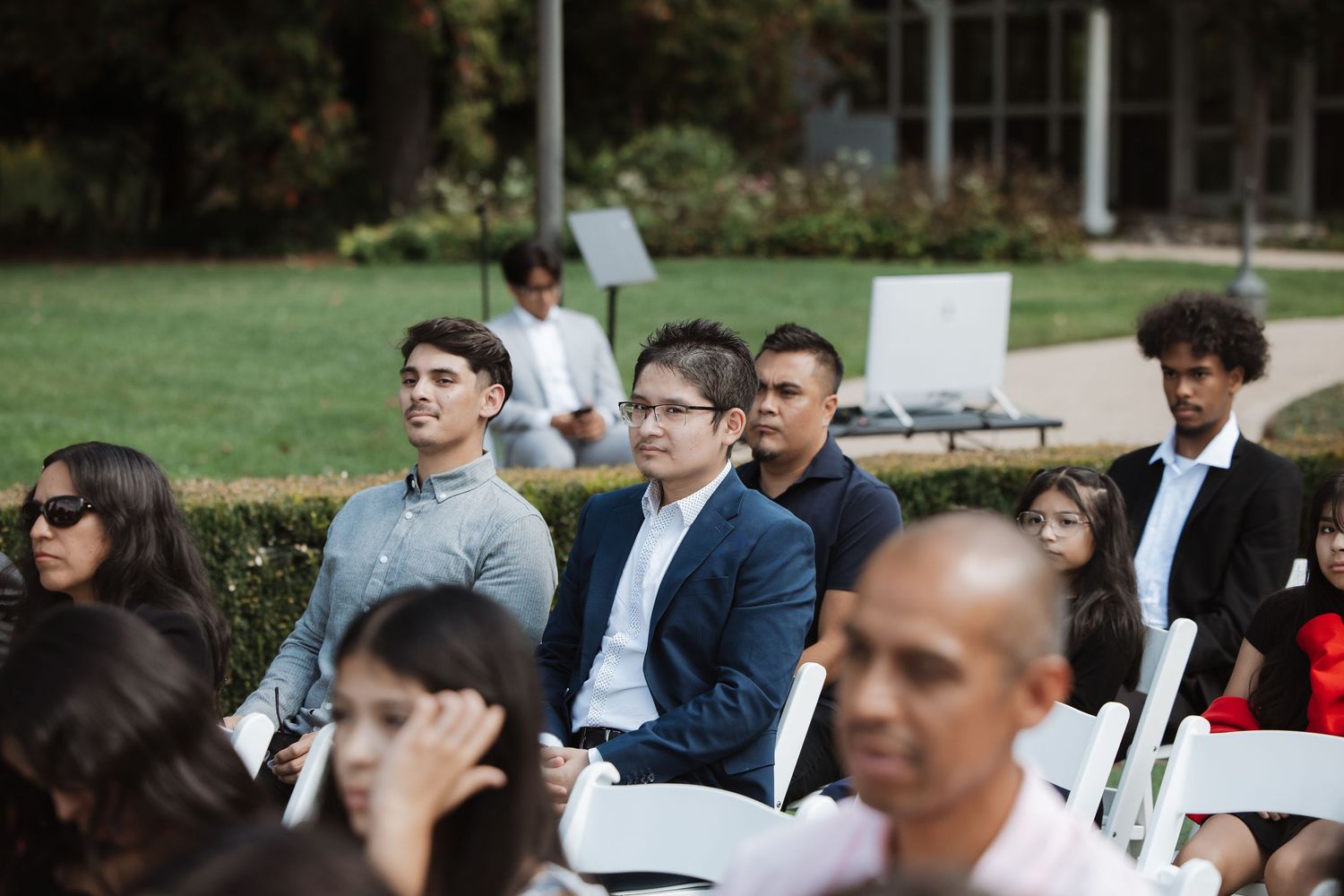 Male wedding guests in casual and formal attire seated at an outdoor garden wedding ceremony.