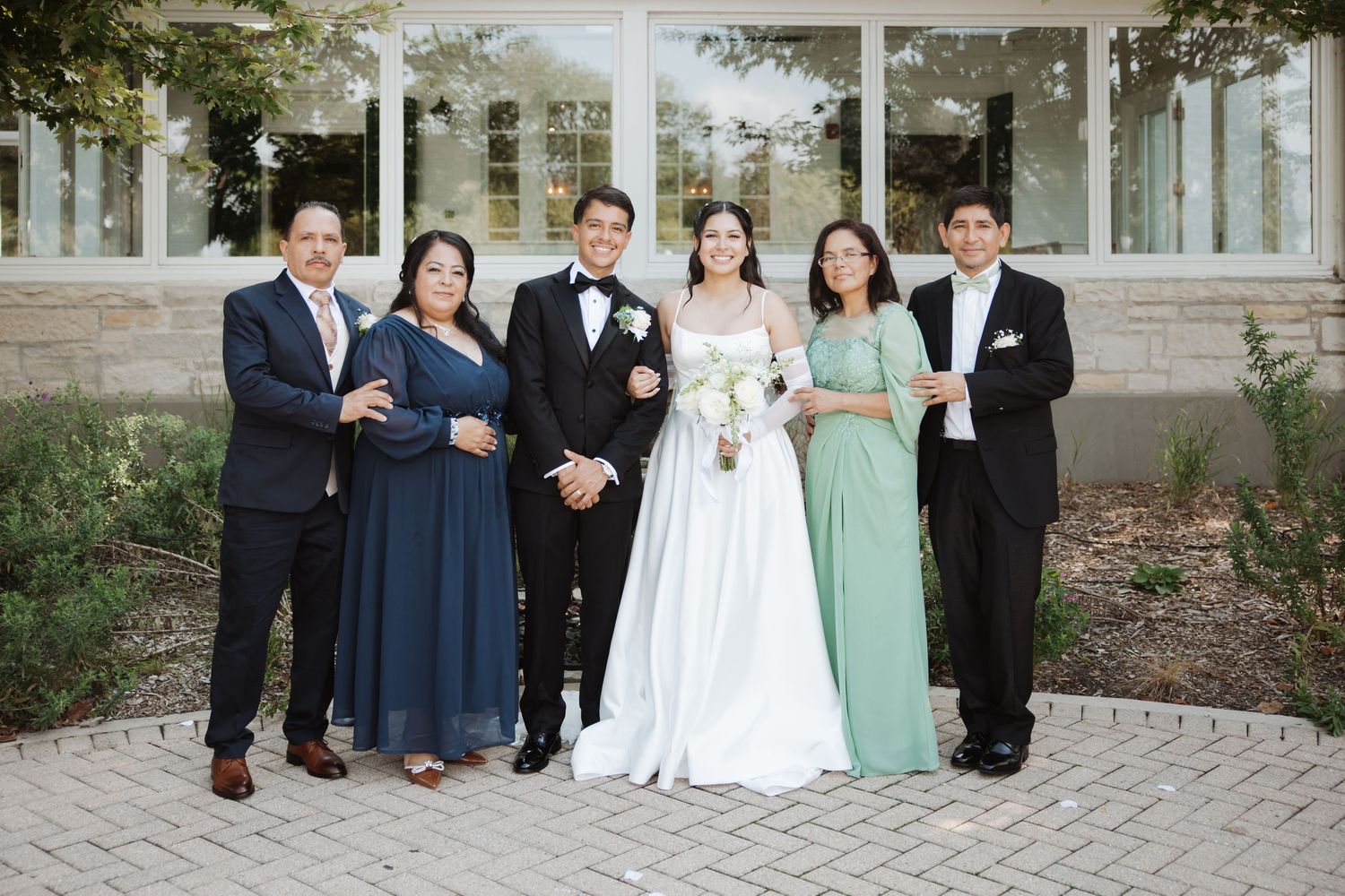 Bride and groom pose formally with parents and sibling in navy and sage green attire outdoors.
