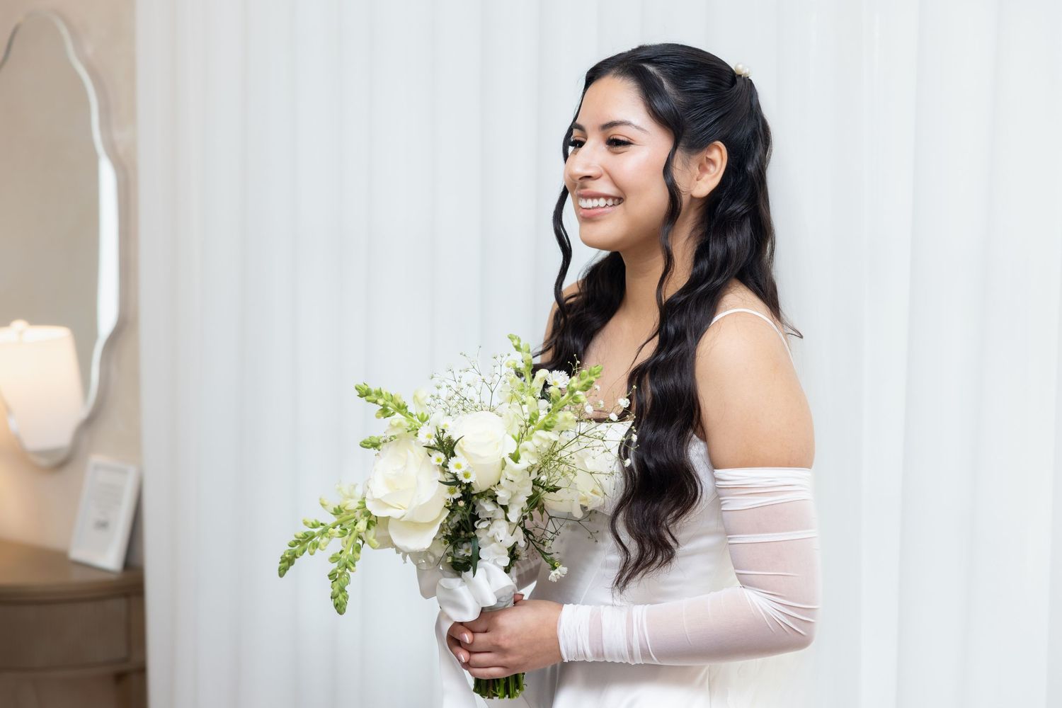 Bride with dark wavy hair in off-shoulder white gown holds white rose bouquet while smiling.