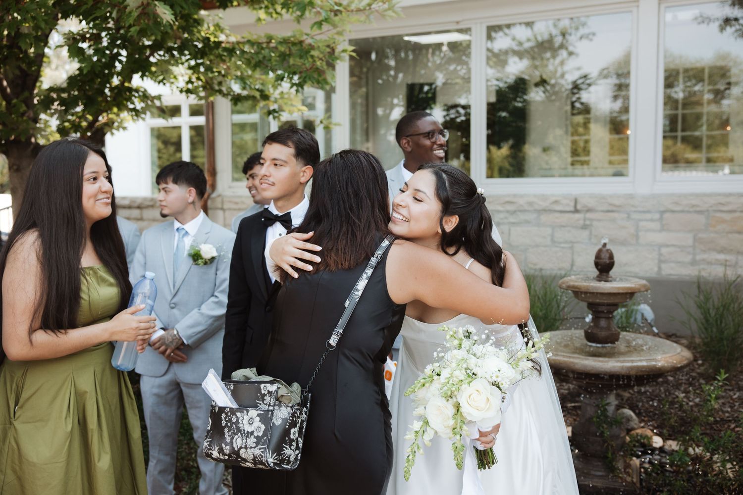 Bride in white gown hugs wedding guest while groom greets others at outdoor post-ceremony celebration.