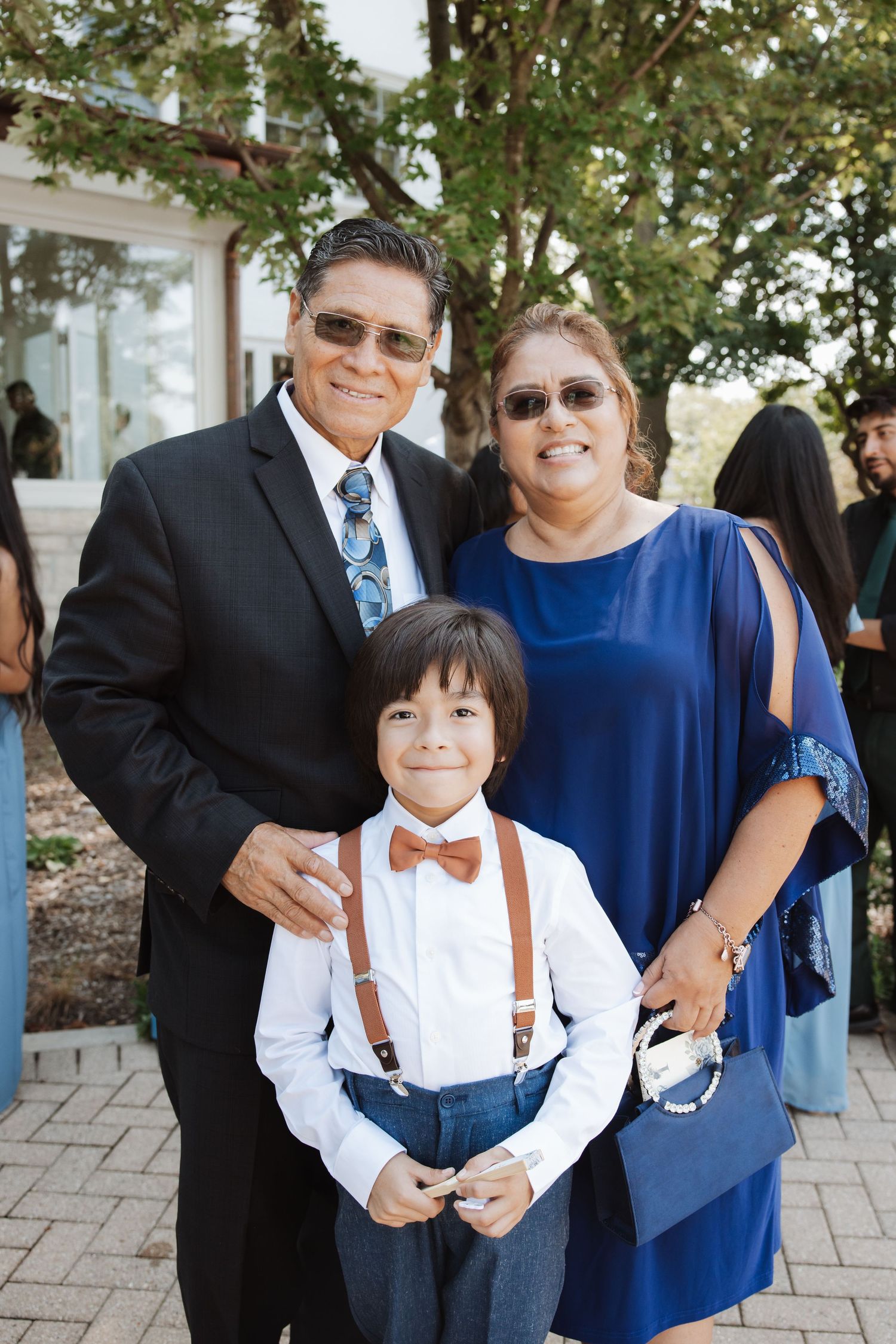 Young ring bearer in suspenders and bow tie poses with grandparents in formal attire at outdoor wedding.