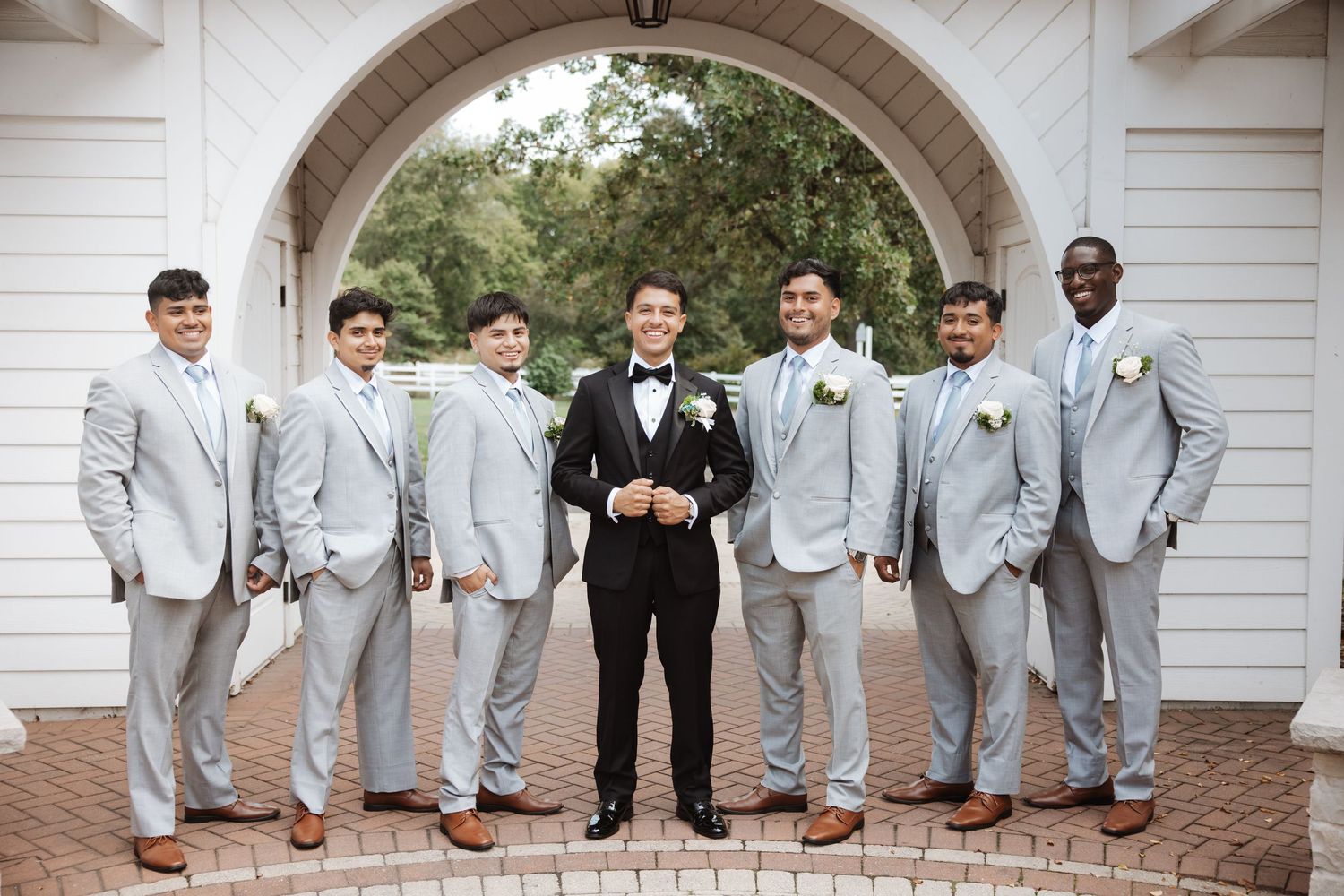 Smiling groom and groomsmen in gray suits pose casually under outdoor arch.