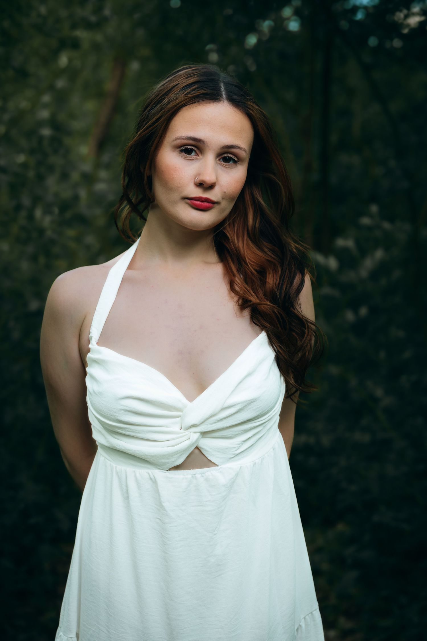 A young woman with long brunette hair wearing a white halter dress poses in front of a dark forest backdrop.