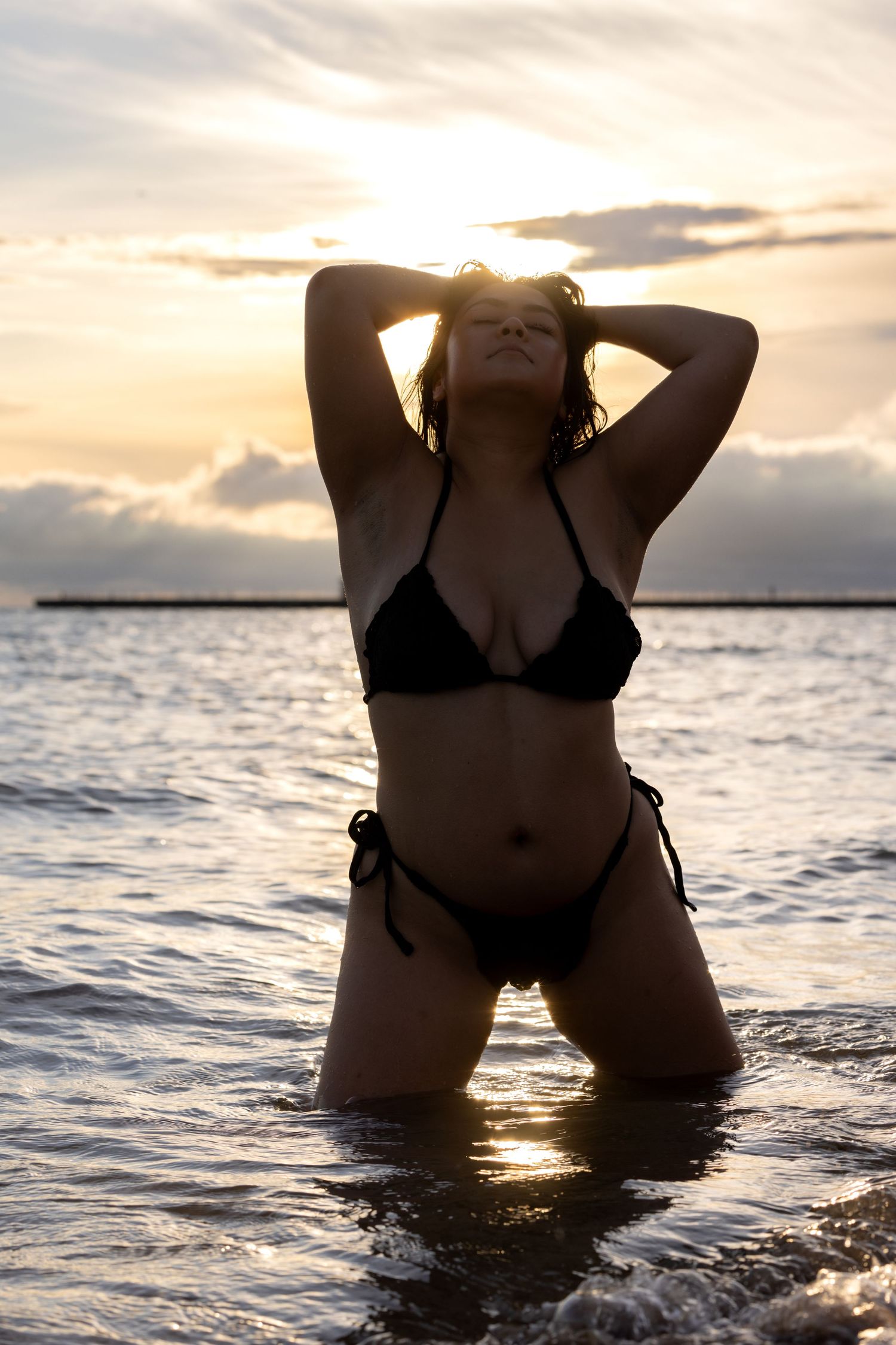 Woman in black bikini kneeling in ocean waves with golden sunset behind her.