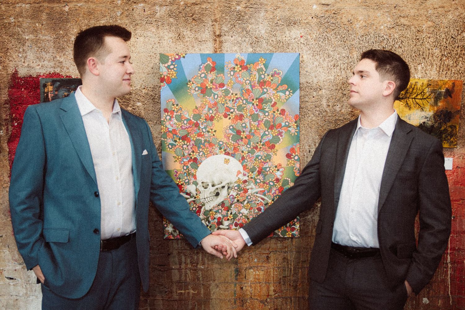 Two men in suits smiling and holding hands in front of a colorful floral skull painting.