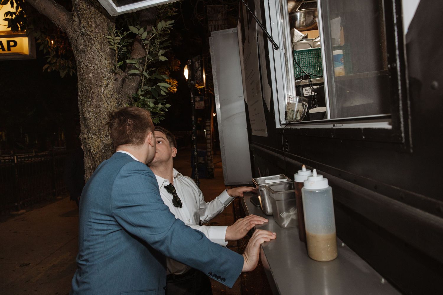 Two men in suits share a kiss at a food truck window during an evening wedding reception.
