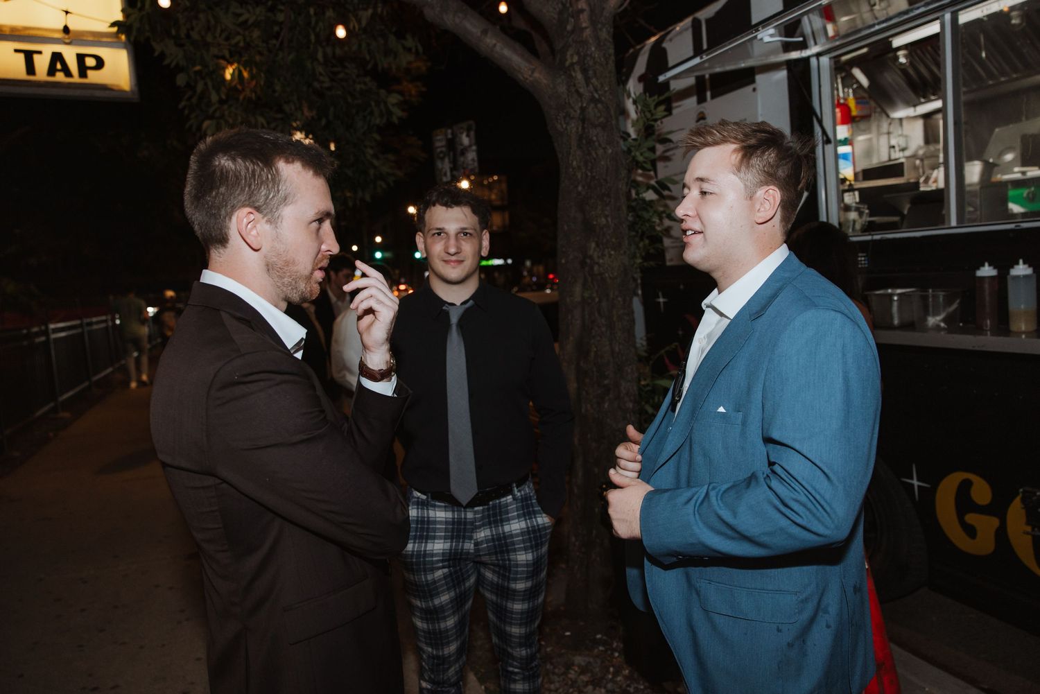 Three men in suits conversing outdoors at night near a tap sign.