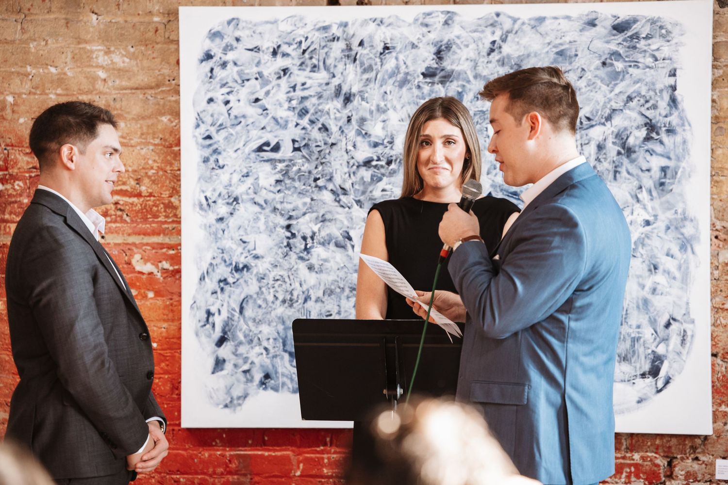 Three people stand before abstract art at a formal ceremony in a brick-walled venue.
