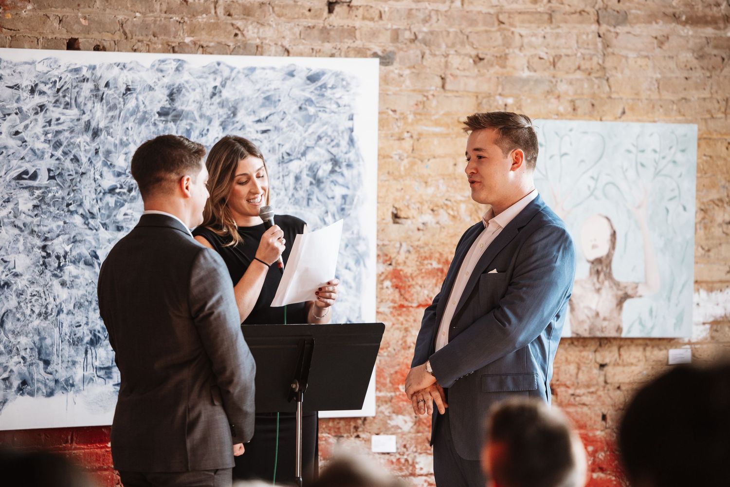 Female officiant reads vows as two grooms stand together during their wedding ceremony in an art gallery.