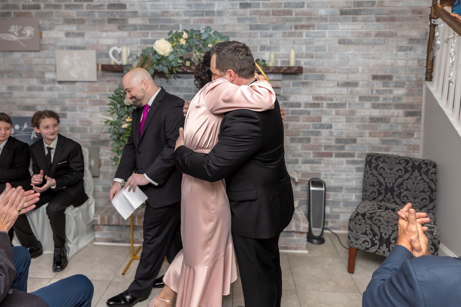 Bride and groom share warm embrace as wedding guests applaud during intimate indoor ceremony.