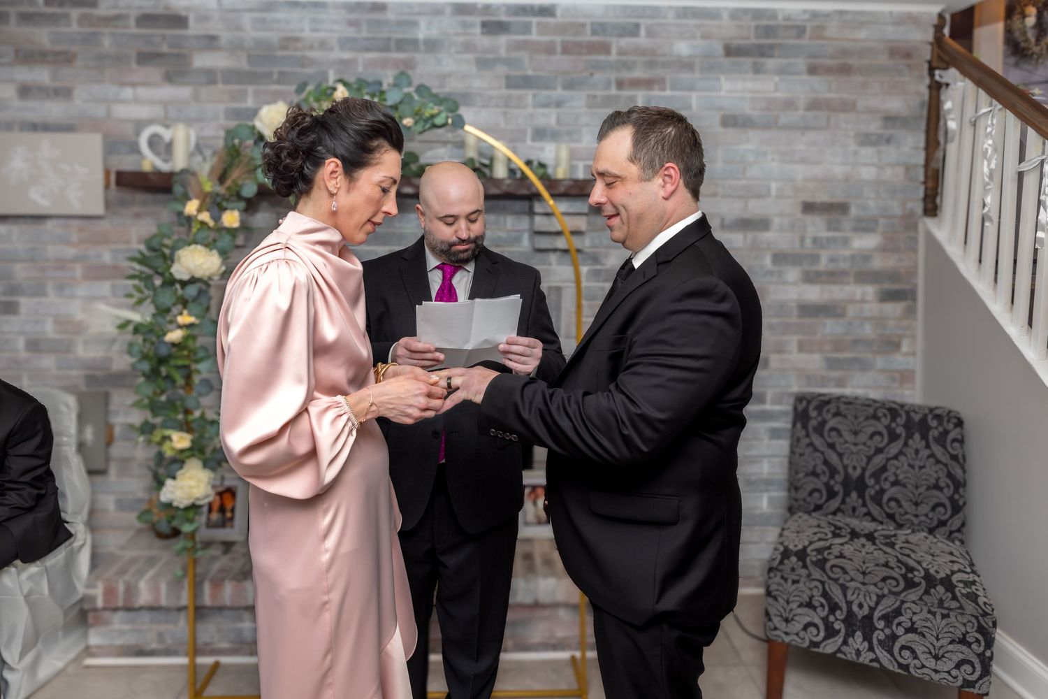 Couple exchanges wedding vows while officiant in purple tie oversees intimate indoor ceremony.