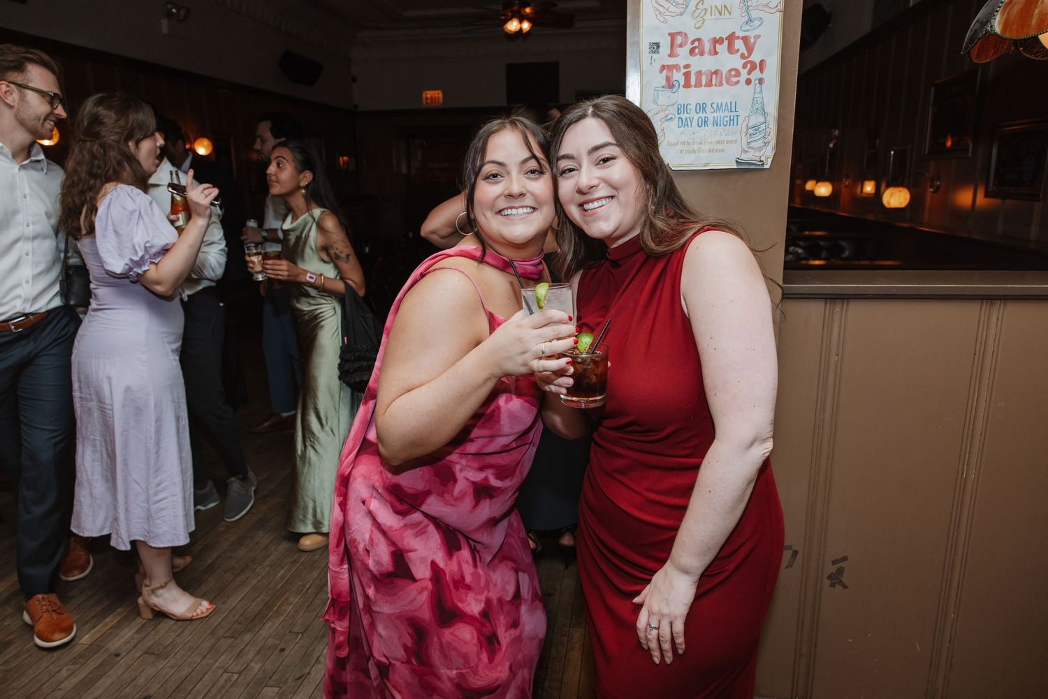 Two women in red and pink dresses smile holding drinks at a party venue.