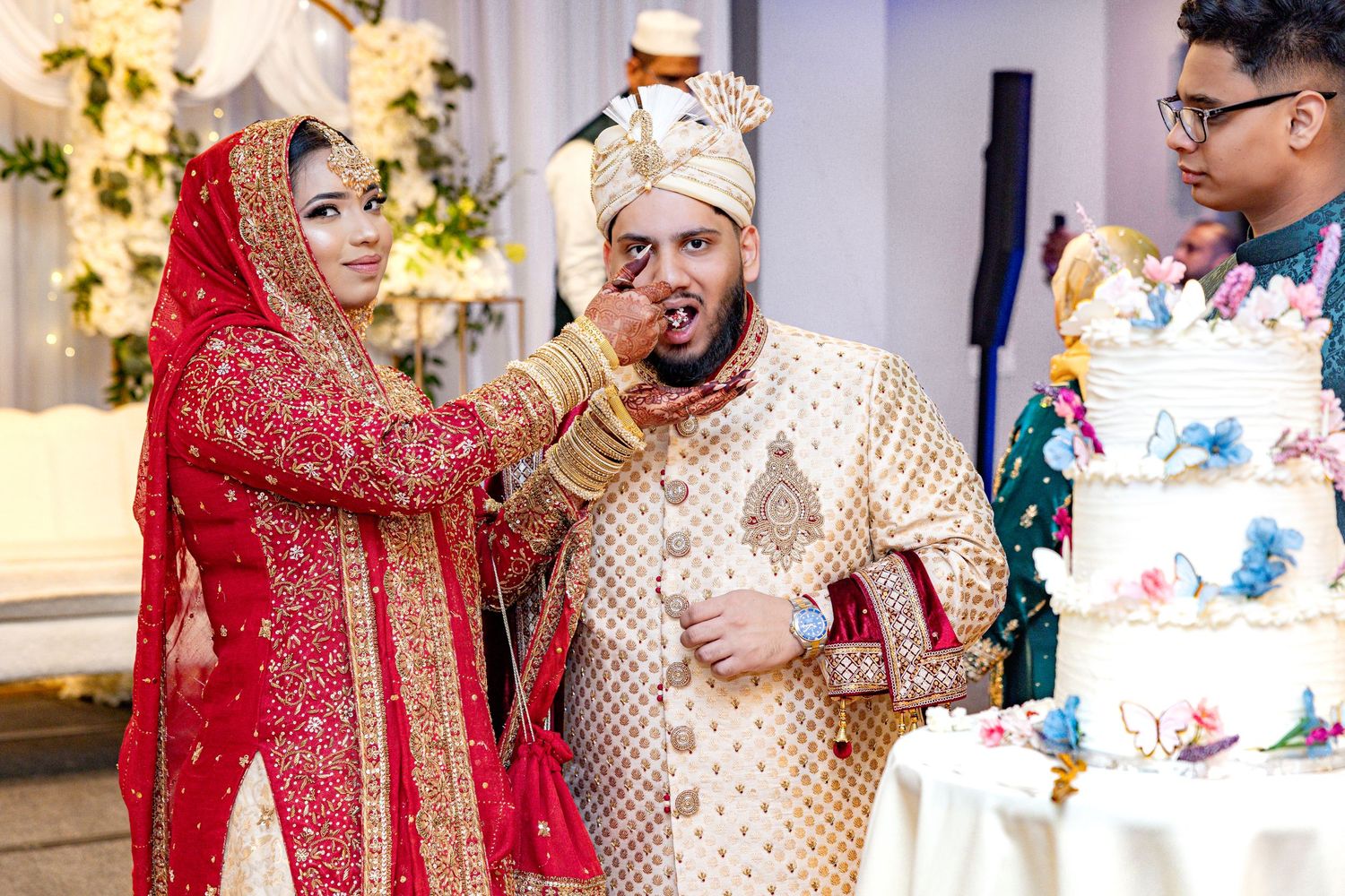 Bride in red lehenga feeding groom cake at traditional South Asian wedding.