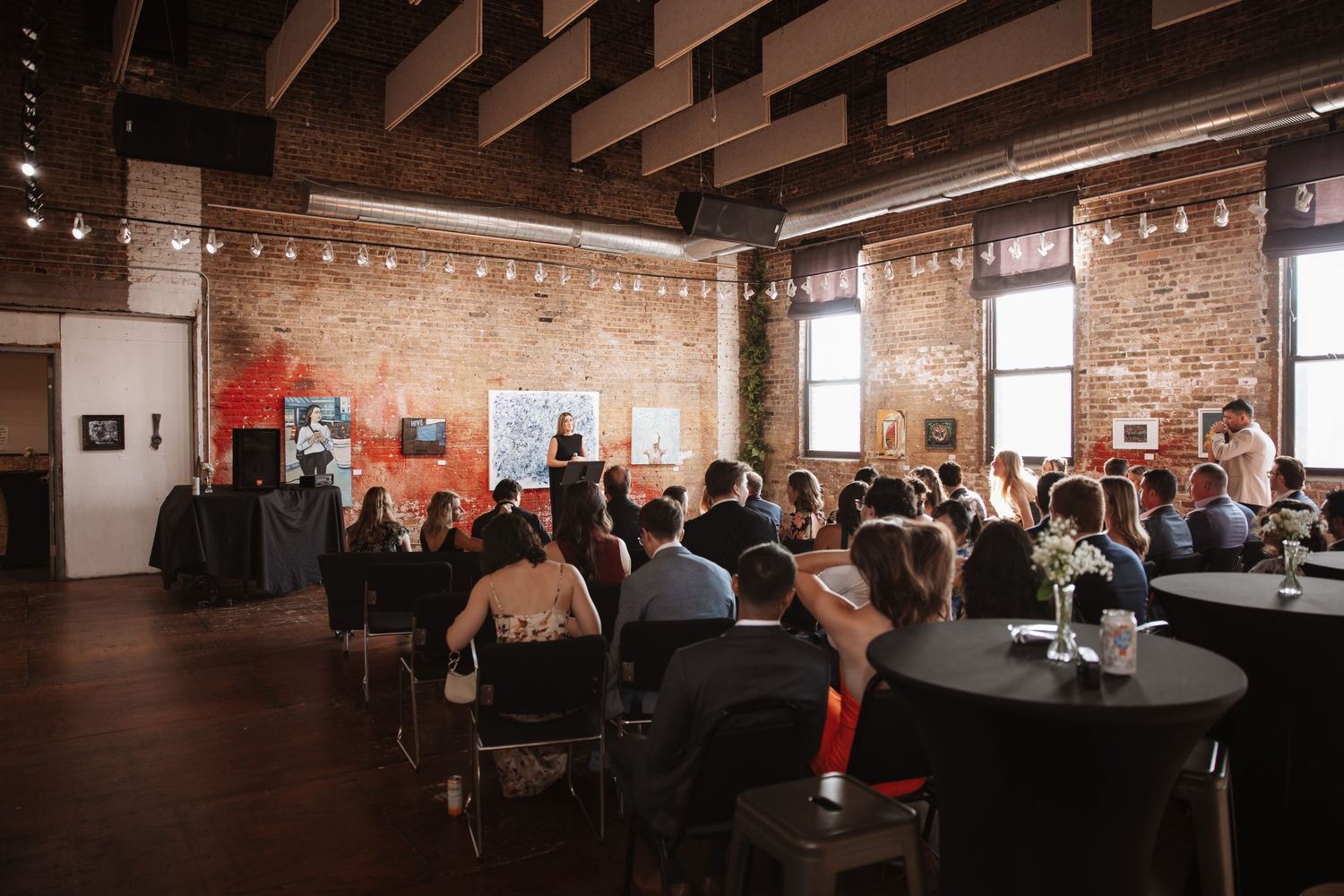 Guests seated and mingling at an indoor event venue with exposed brick walls and string lights.