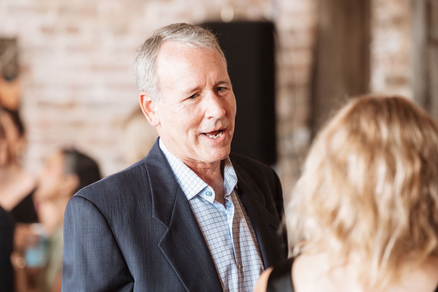 Older man in a navy blazer smiling warmly while conversing at an indoor event.