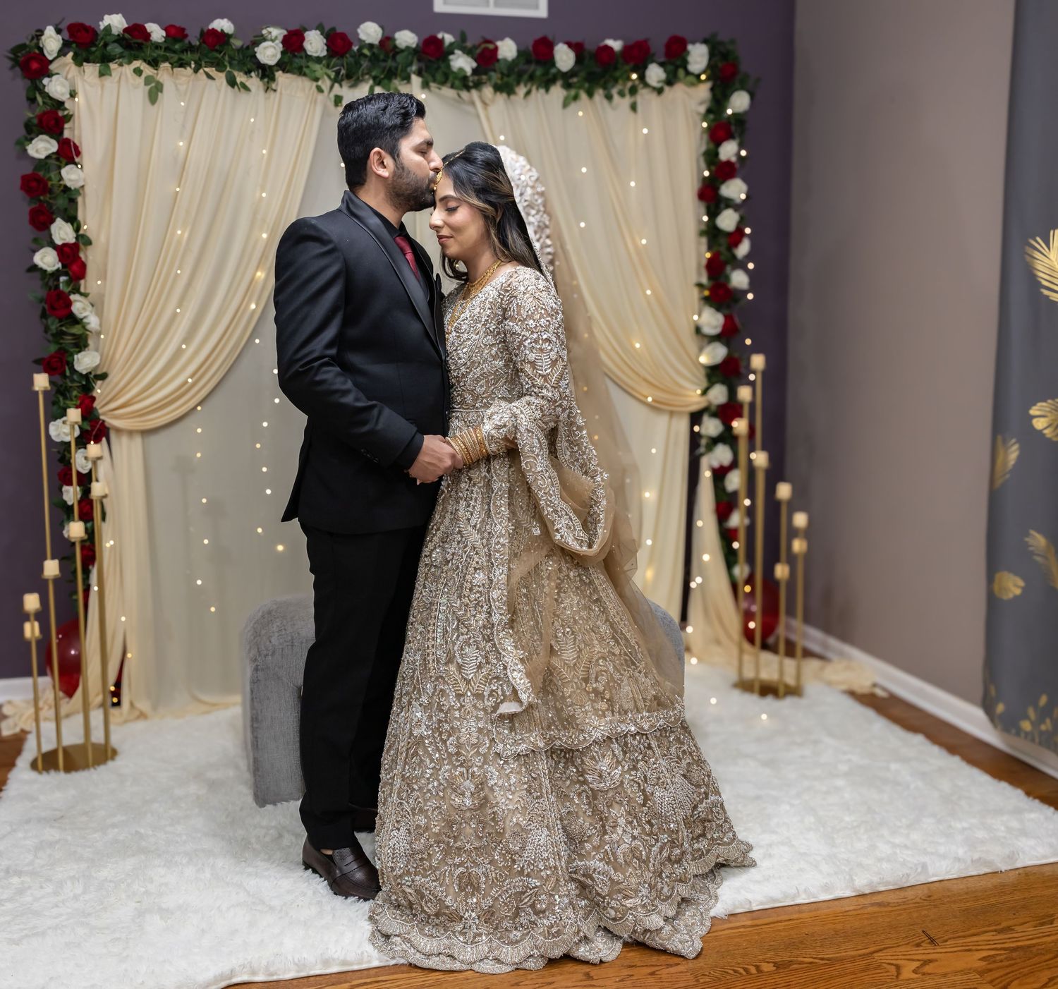 Bride and groom in elegant attire share a romantic embrace before cream and floral wedding backdrop.