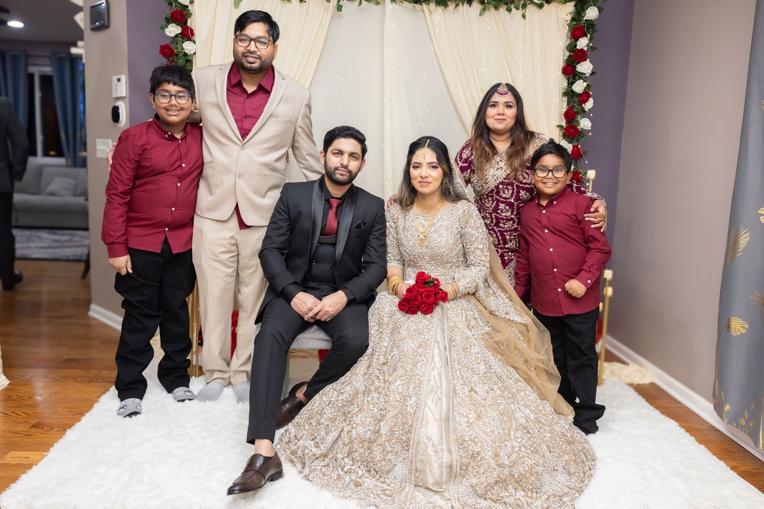 South Asian wedding couple with family posing at decorated mandap with floral arrangement.