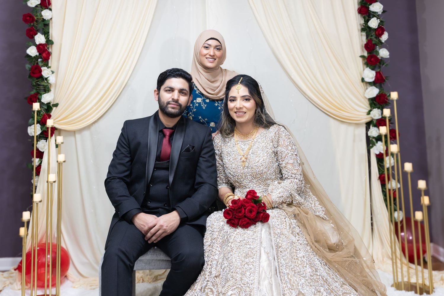 South Asian bride and groom with female guest posing at floral decorated wedding mandap.
