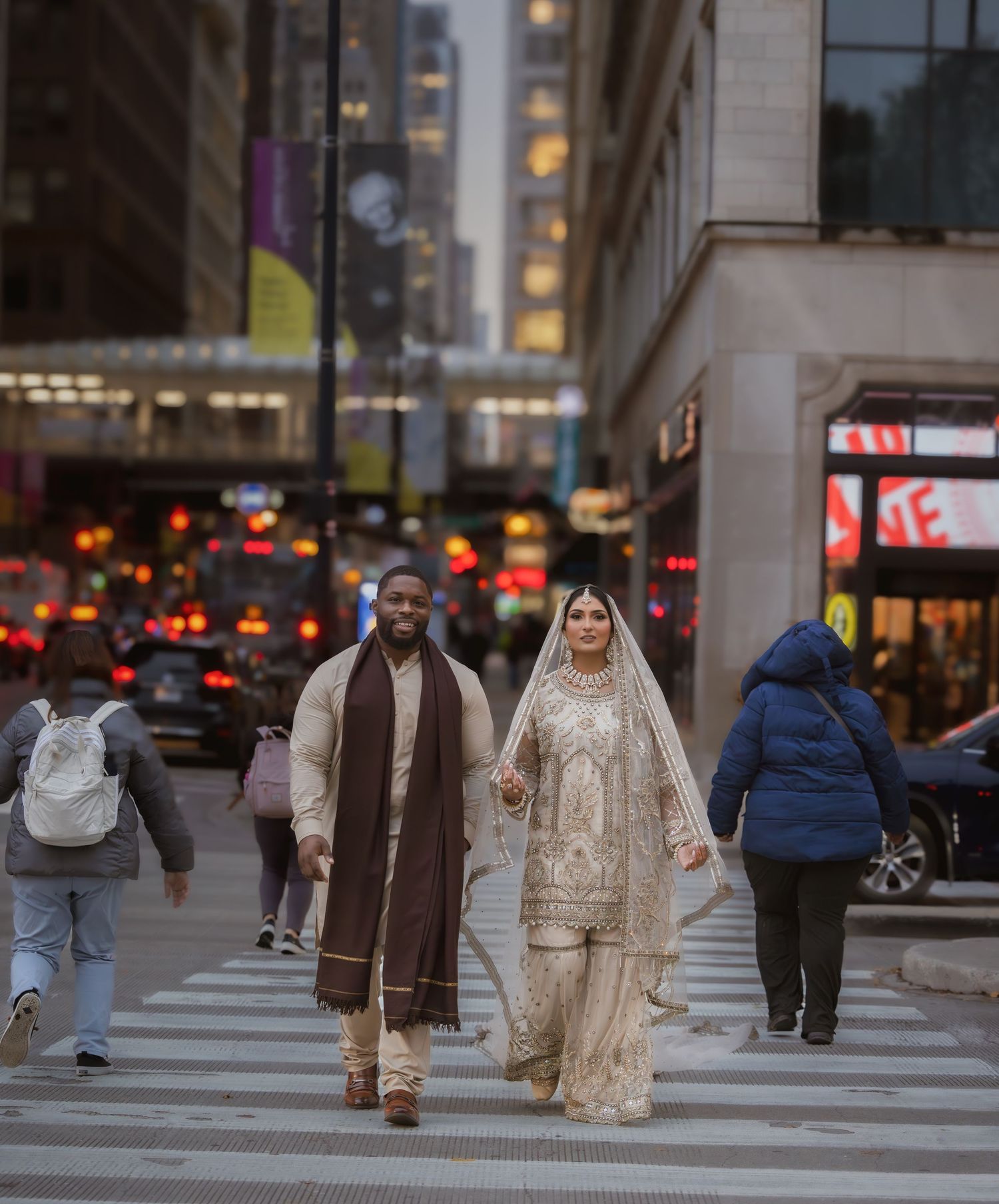 Interracial wedding couple in traditional South Asian attire crossing a busy city street at twilight.