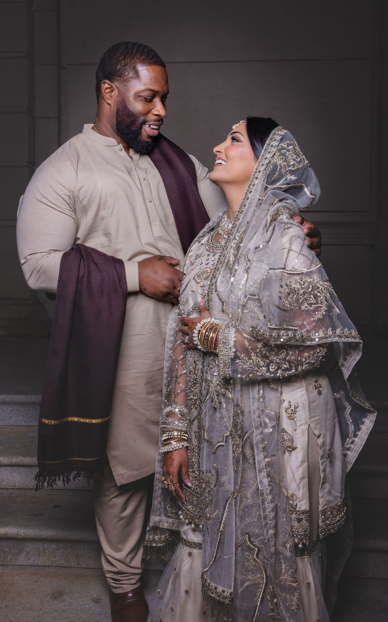 Interracial Muslim couple in traditional wedding attire smiling at each other on marble steps.