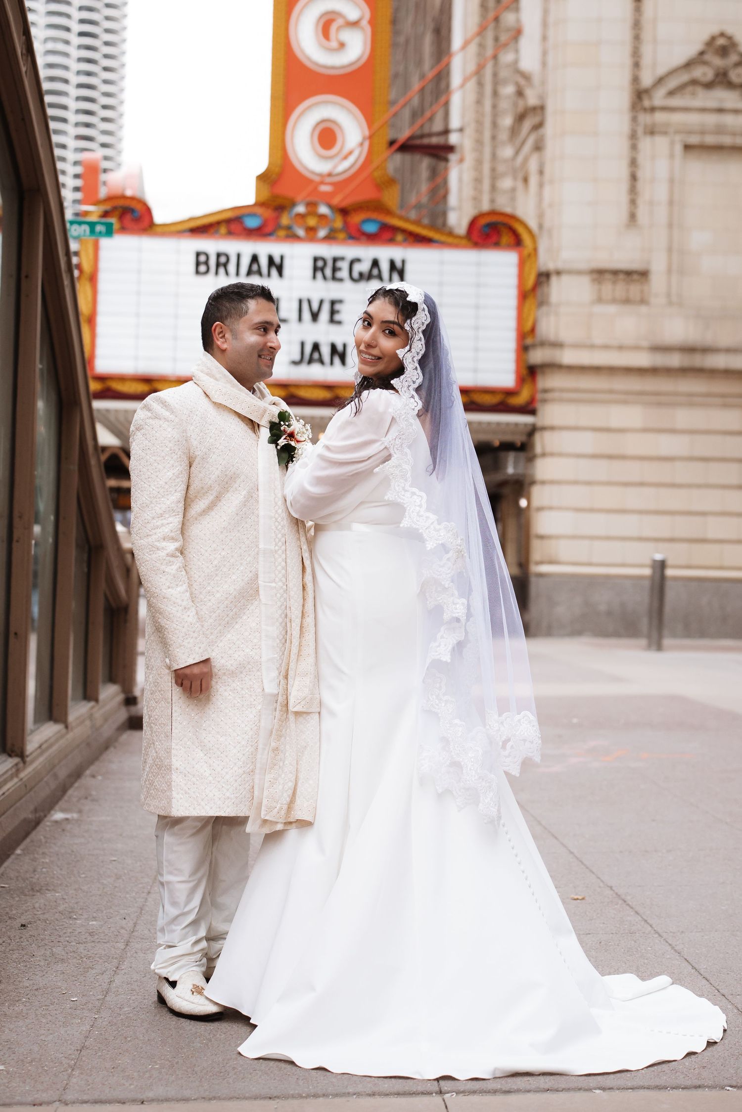 South Asian bride in lace veil and groom in cream sherwani pose full-length outside Chicago Theatre.