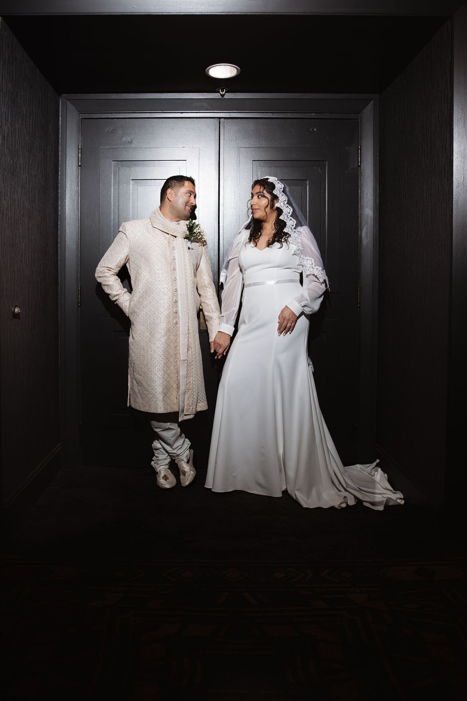 Bride in white gown and veil holds hands with groom in cream sherwani standing before dark doors.