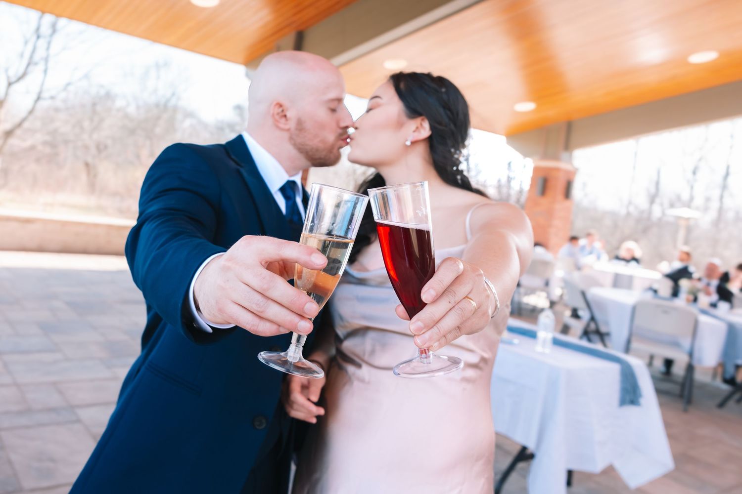 A newlywed couple kisses while clinking champagne and red wine glasses at their wedding reception.