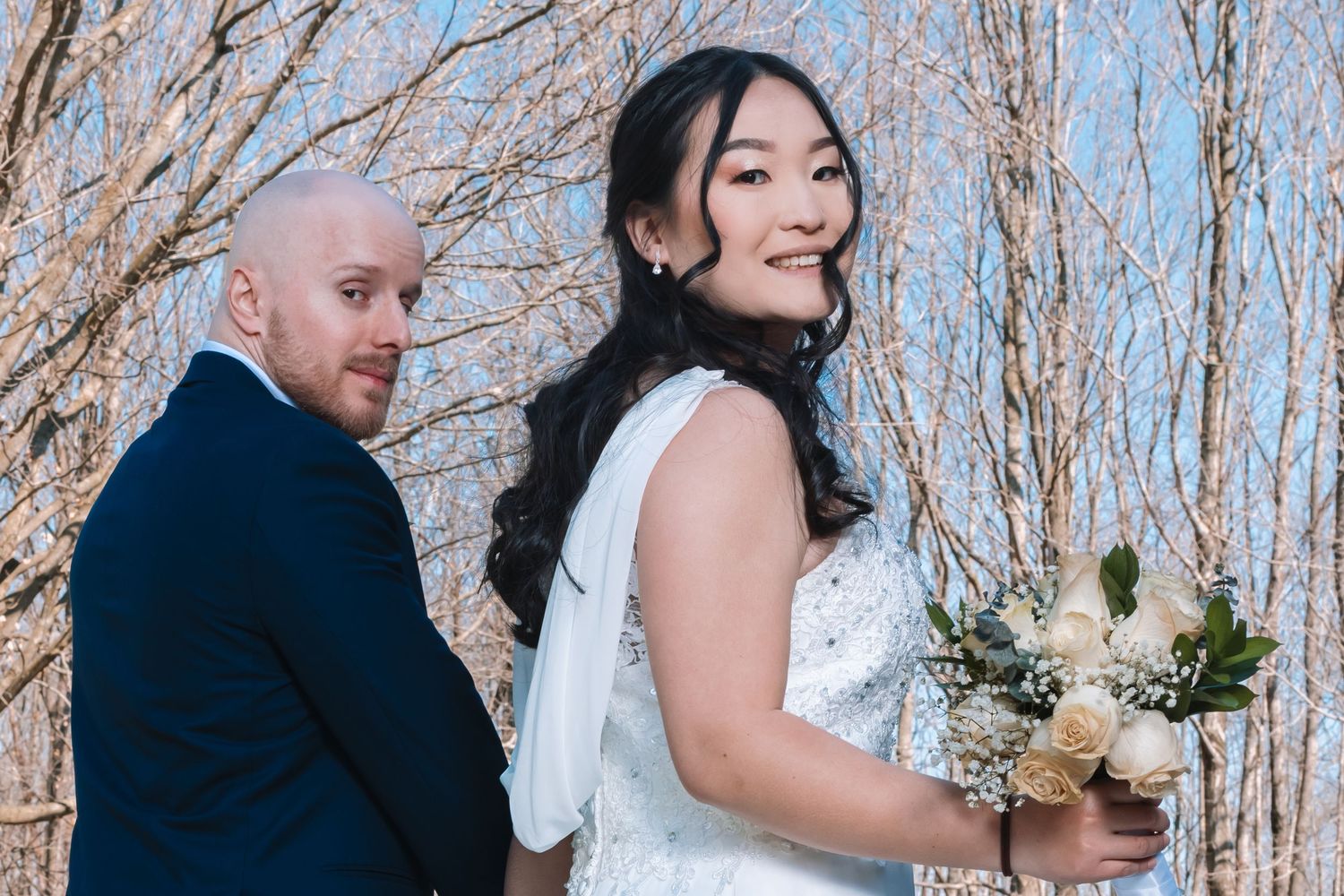 Bride holding bouquet glancing back at groom among bare winter trees.