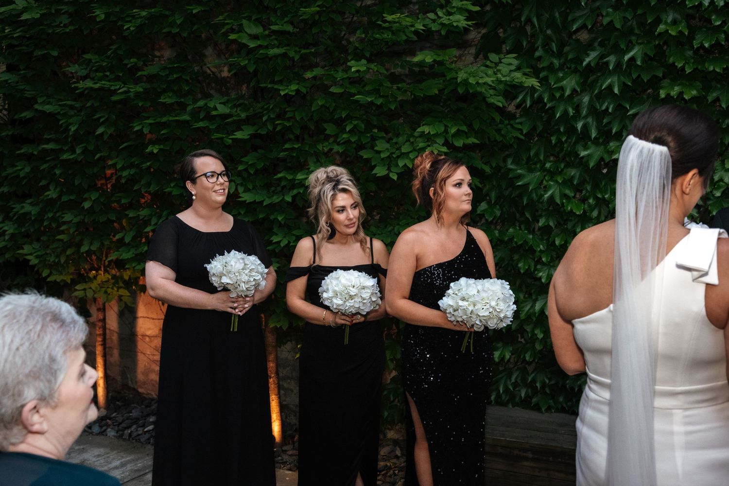 Three bridesmaids in black dresses hold white bouquets during an outdoor wedding ceremony.