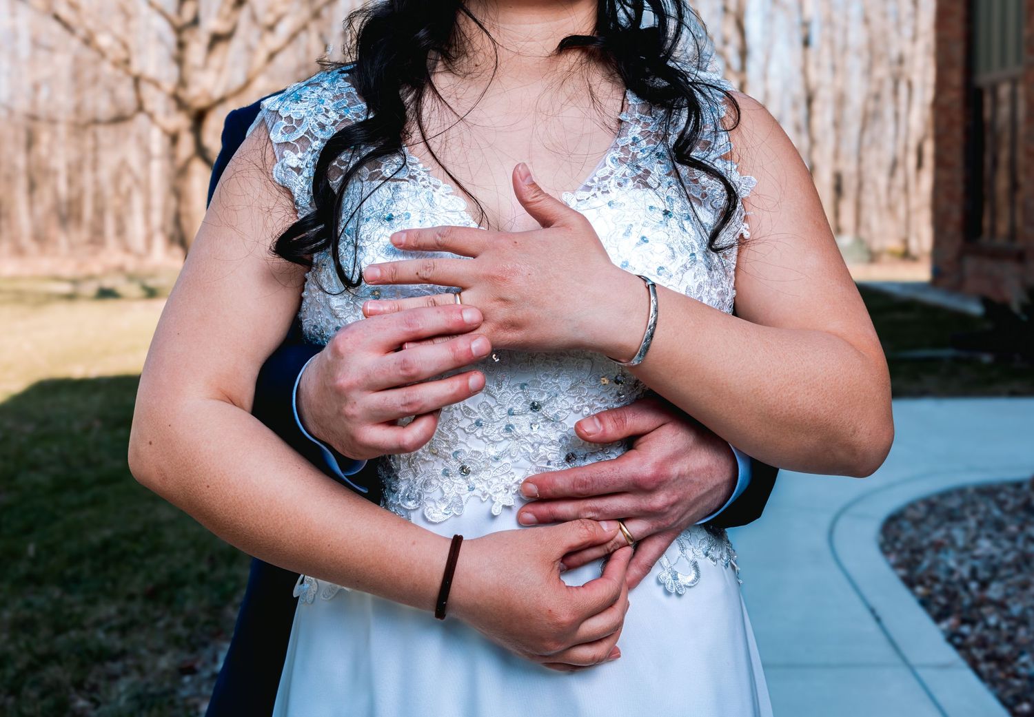 Groom embracing bride from behind showing wedding rings and bracelets in outdoor wedding portrait.
