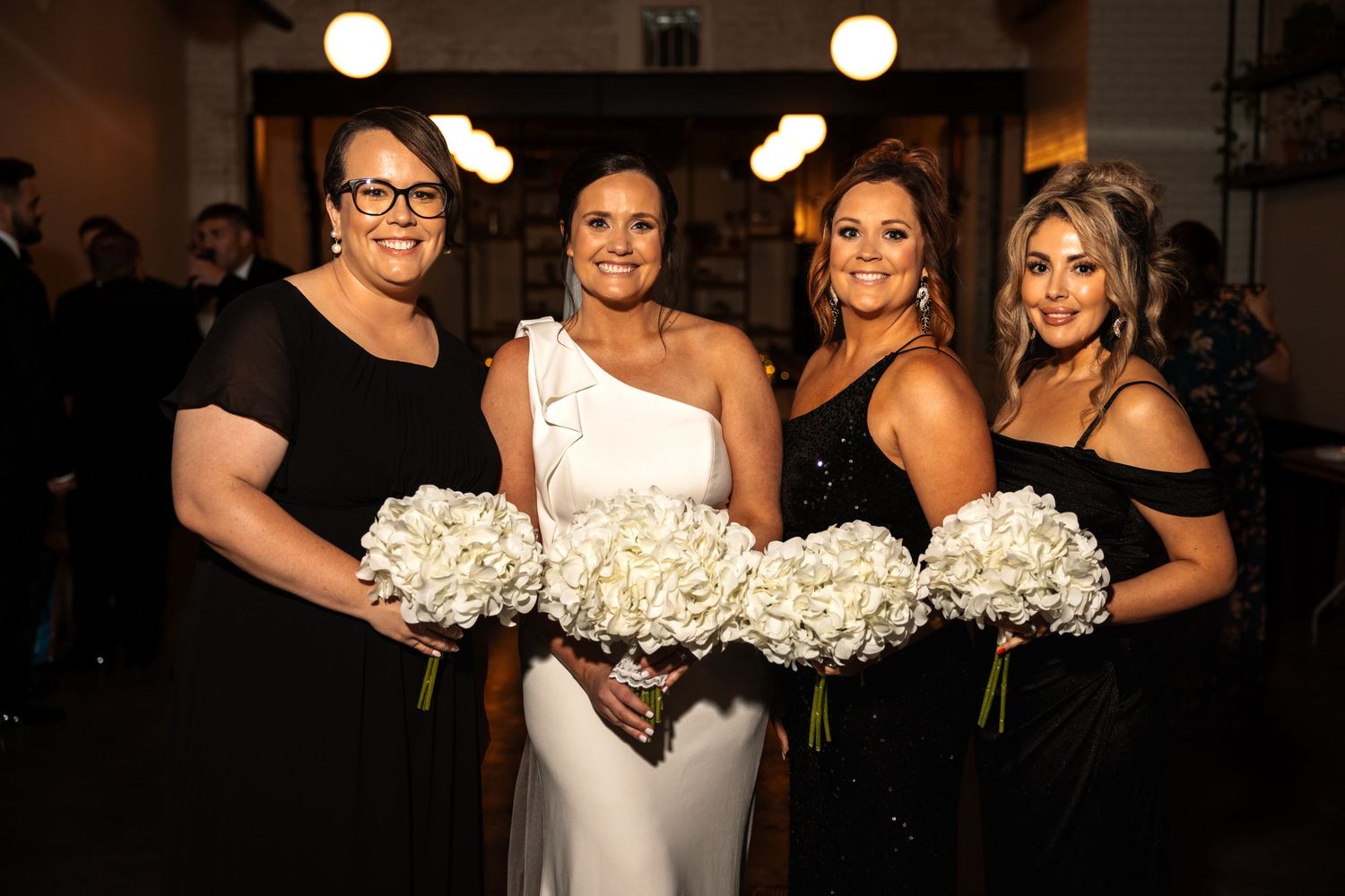 Smiling bride with three bridesmaids in black dresses displaying large white hydrangea bouquets at wedding.