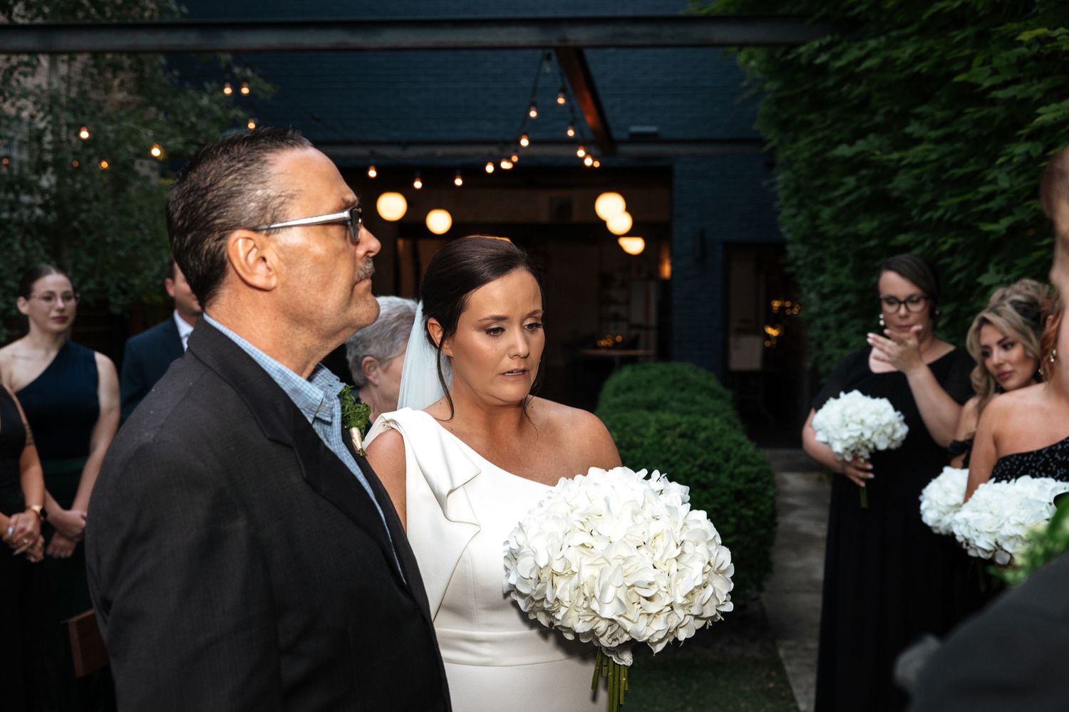 Bride in white off-shoulder gown holding white hydrangea bouquet walking with escort at outdoor wedding.