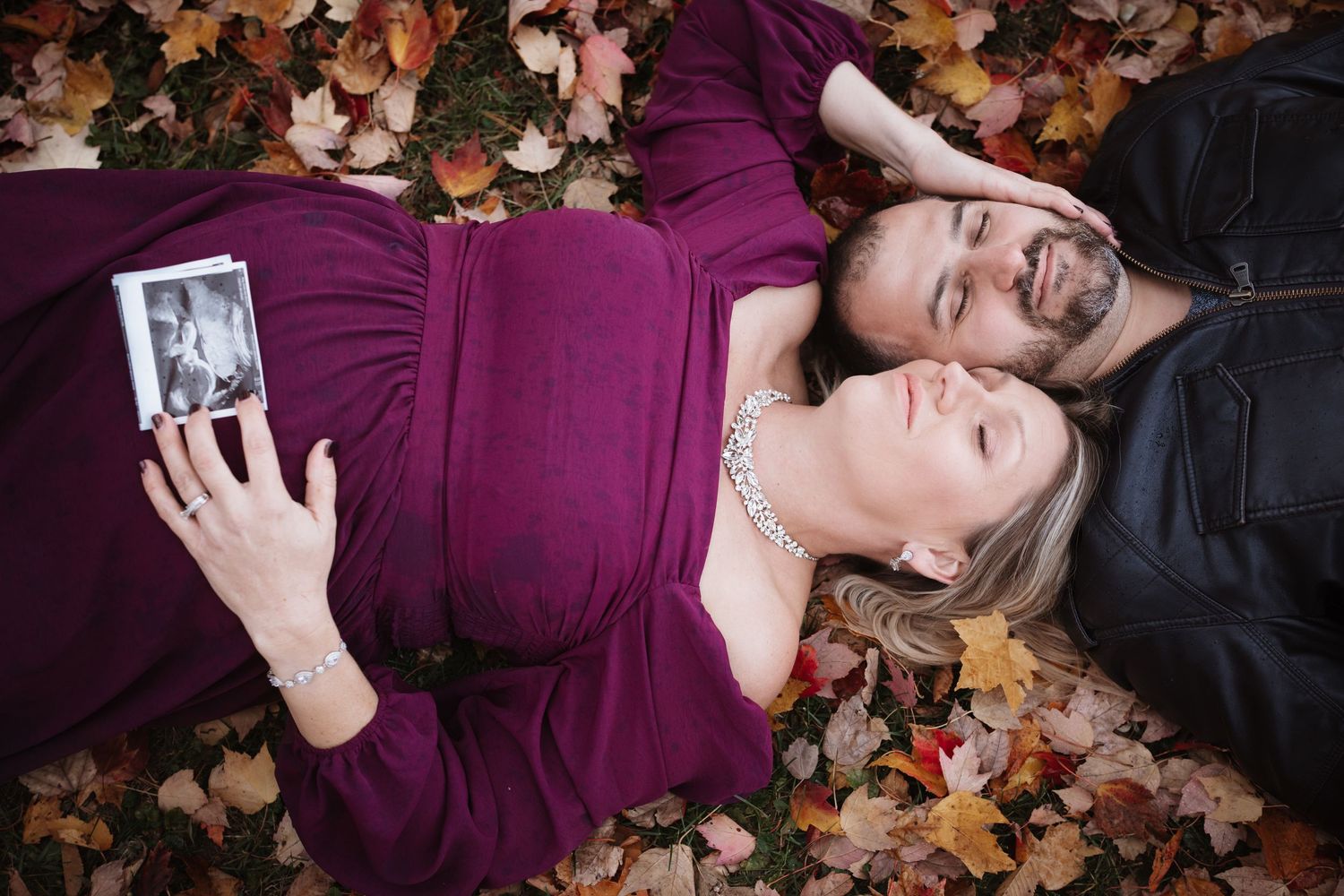 Couple lying in fall leaves, pregnant woman in purple dress holding ultrasound photo, partner beside her.