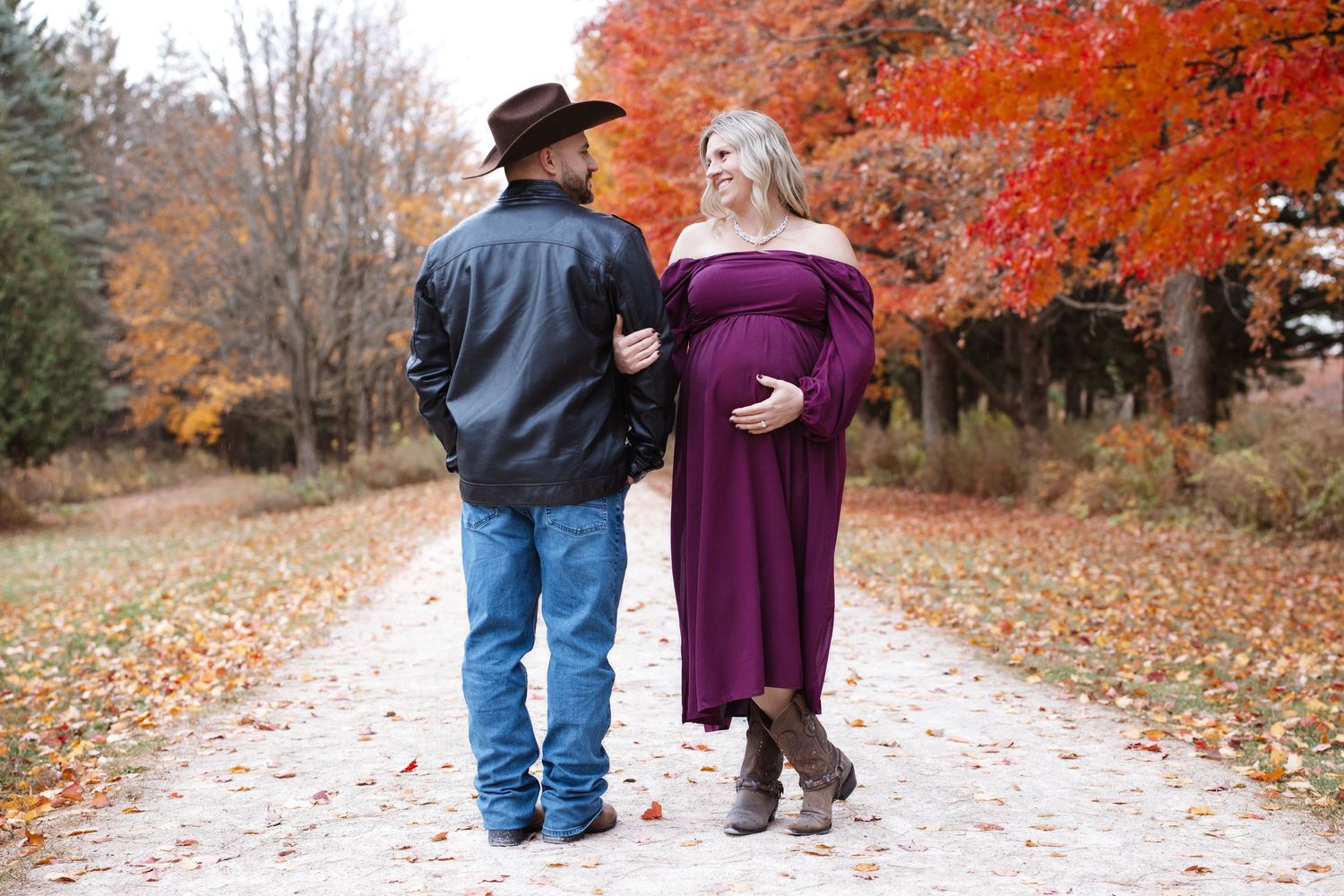 Pregnant woman in purple maternity dress smiling at partner in cowboy hat on fall foliage path.