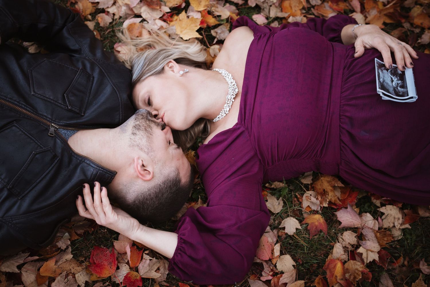 Couple sharing a kiss while lying in autumn leaves, pregnant woman in purple dress holding ultrasound photo.