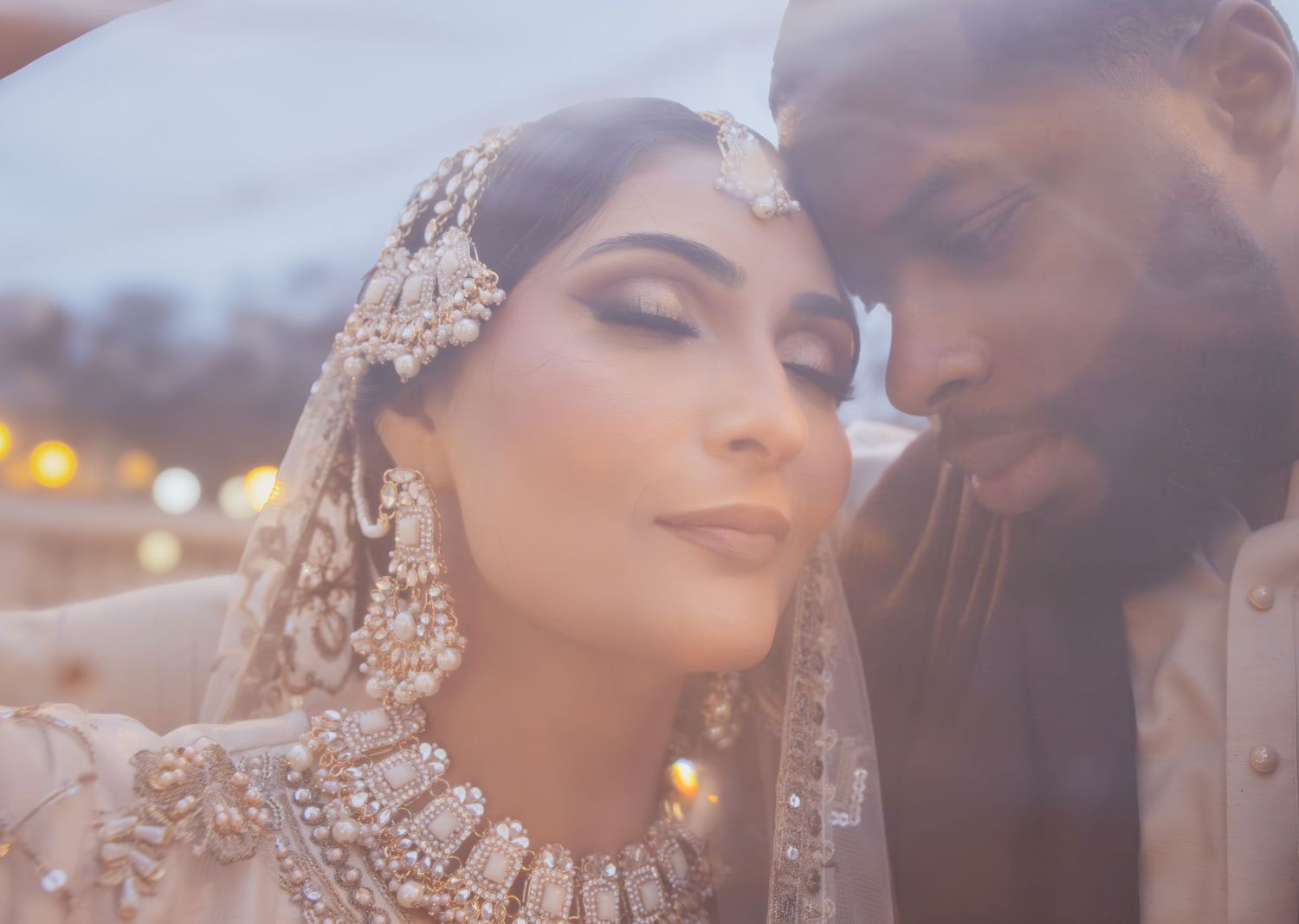 South Asian bride in gold bridal jewelry and lehenga leans close to groom in cream sherwani at dusk.