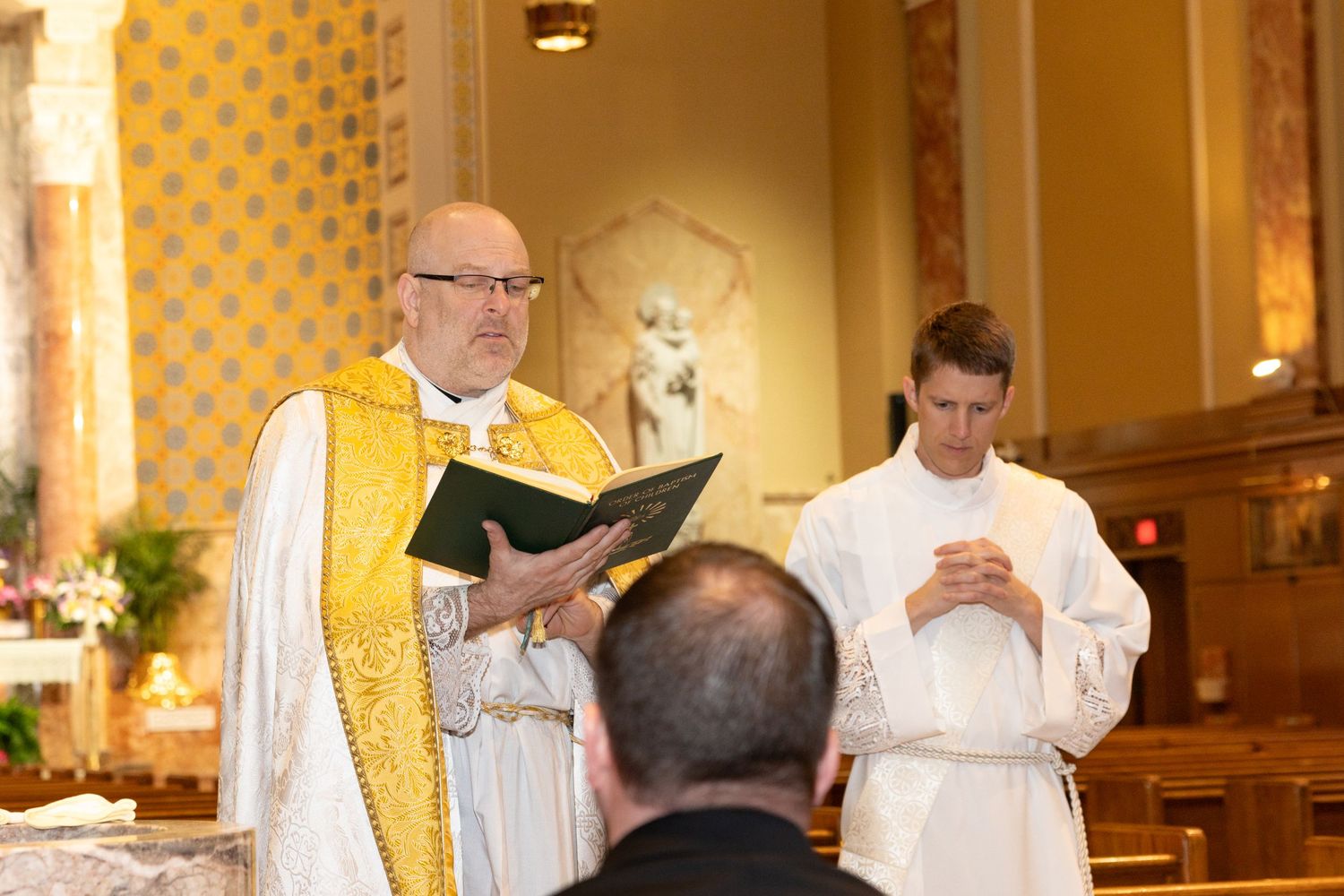 Catholic priest in gold vestments reads from a book during a baptism ceremony at the altar.