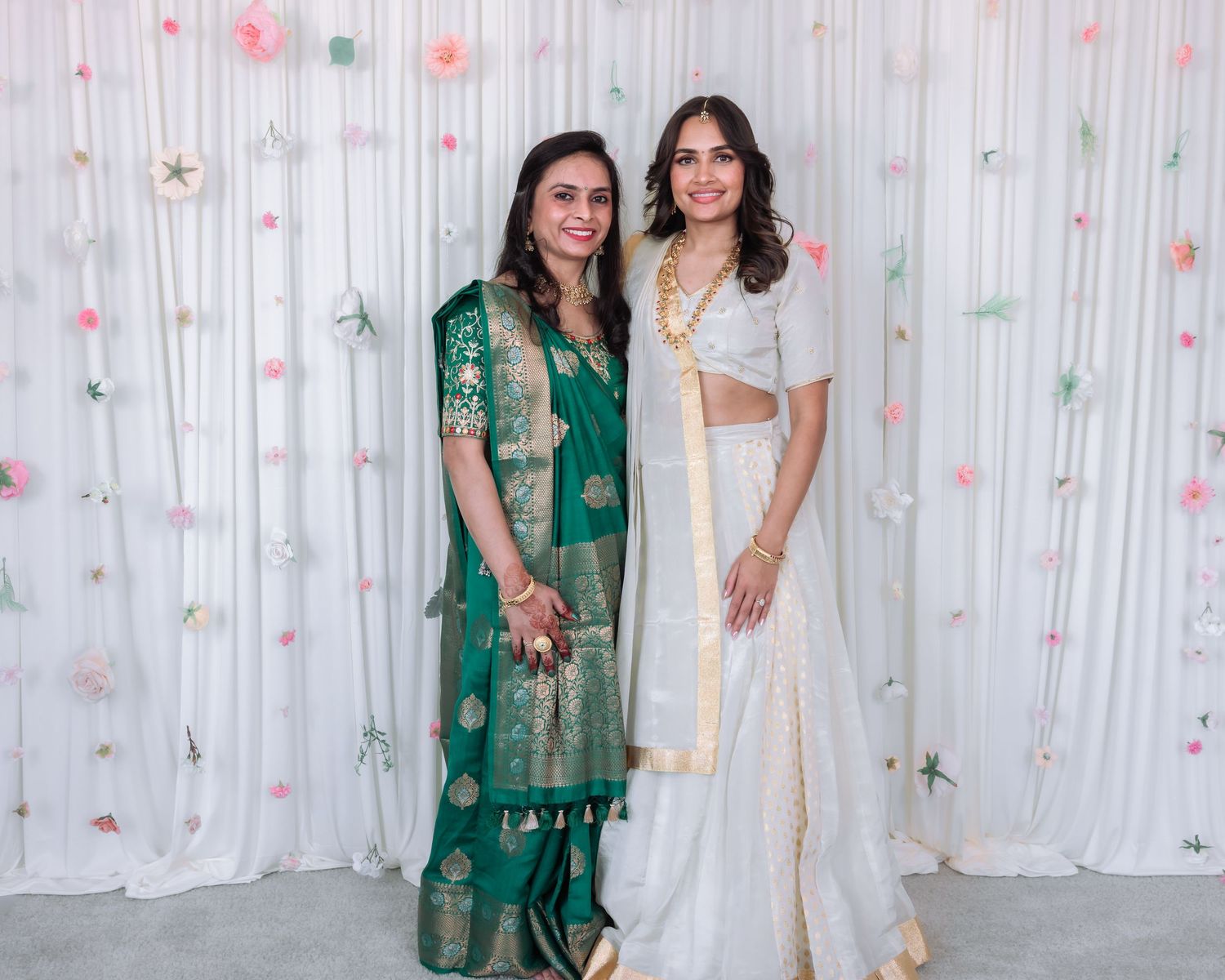 Two women posing together at Indian wedding, one in green saree and one in white lehenga with gold jewelry.