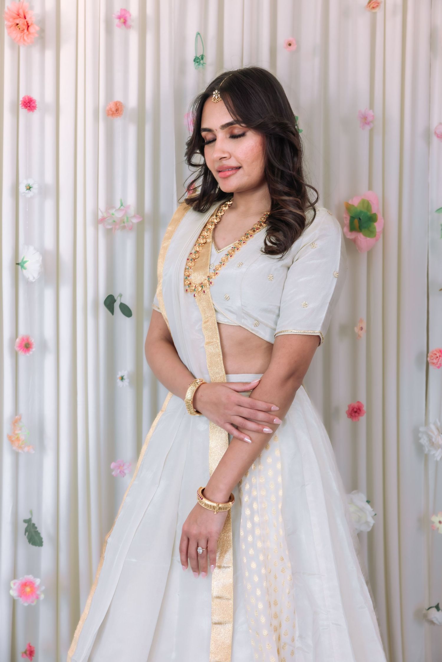 Woman in white gold-trimmed lehenga with eyes closed posing serenely before a floral backdrop.