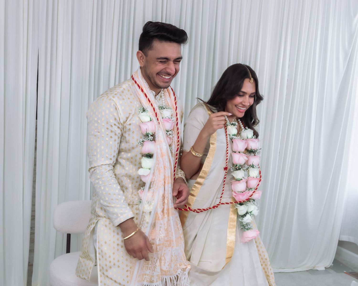 Smiling Indian bride and groom exchange floral garlands in jaimala ceremony wearing white wedding attire.