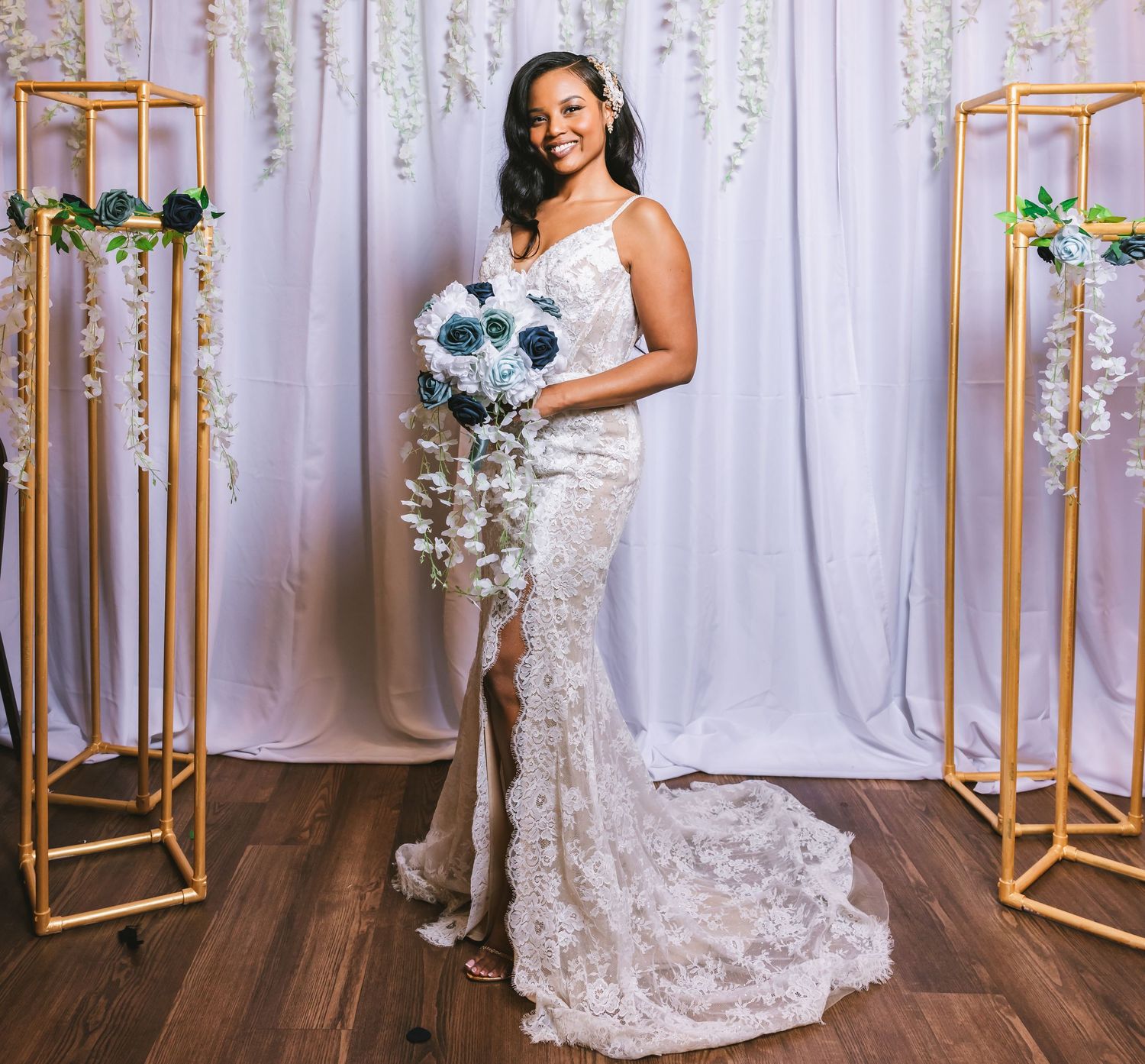 Elegant bride in lace mermaid wedding gown holding blue and white bouquet against white backdrop.