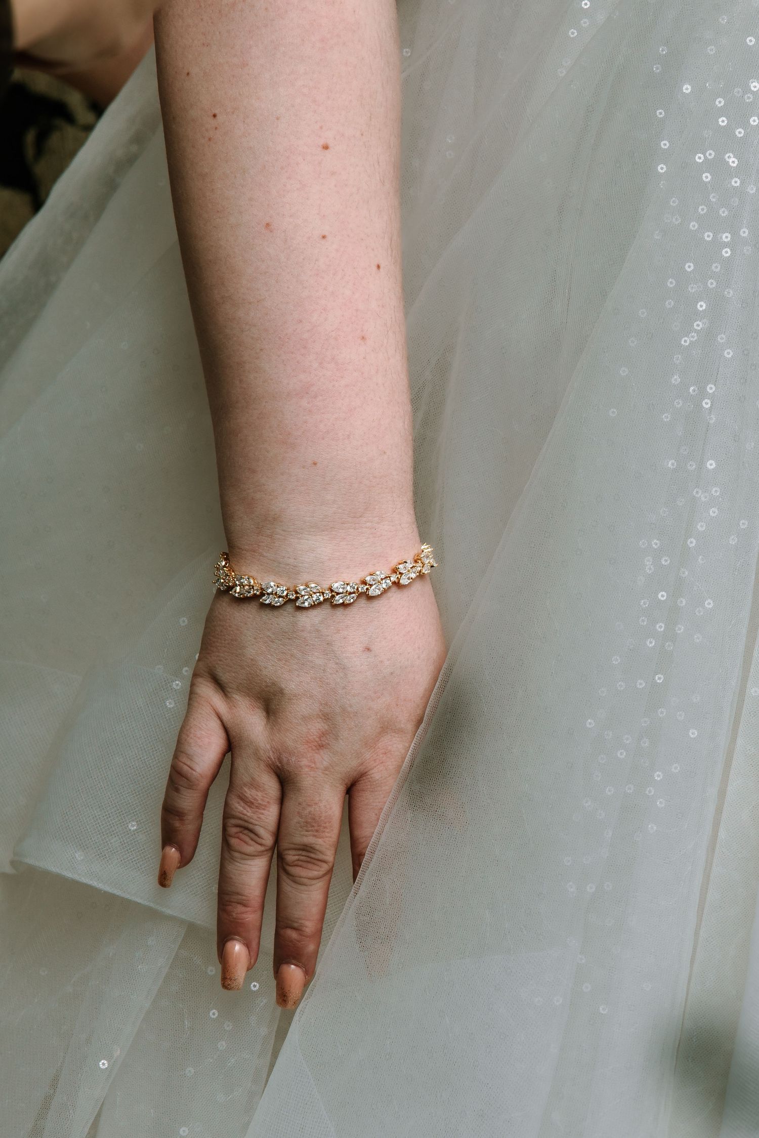 Bride's hand wearing a gold leaf bracelet resting on a sparkly white tulle wedding dress.